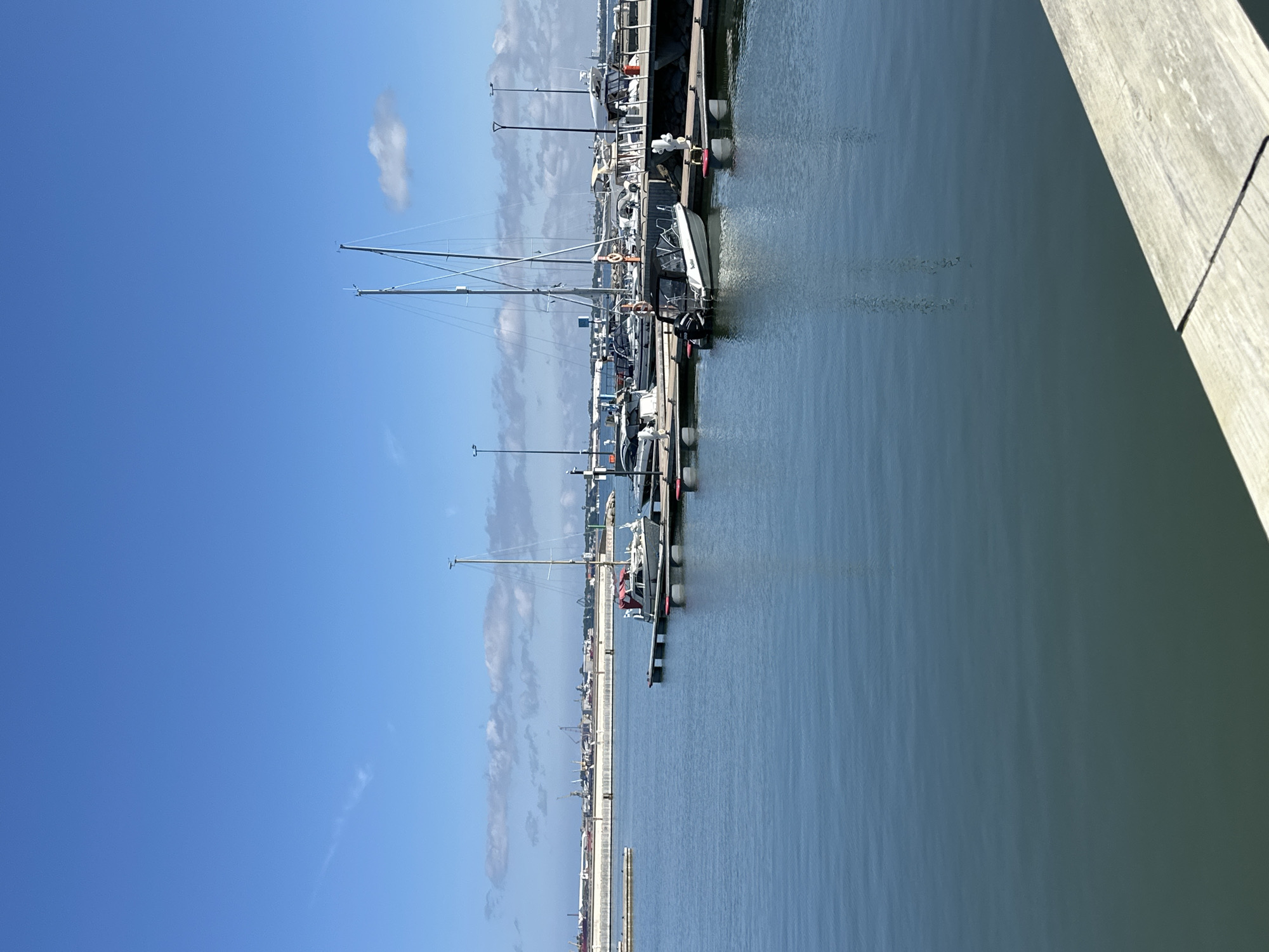 This is a clear, sunny day photo taken at a marina in Tallinn, Estonia, likely in the summer. The image captures a serene view of the harbor with several boats docked. The foreground shows calm, blue water reflecting the sky, with a wooden platform edge in the lower right corner. In the middle ground, a marina with multiple boats is visible. One sailboat with tall masts stands out, along with several smaller motorboats. These boats are moored to a pier. In the background, the cityscape of Tallinn is visible, characterized by a long, white barrier wall, industrial structures, and several cranes, indicating a port area. The sky is a bright, clear blue with a few wispy clouds. There are no people clearly identifiable in the scene.