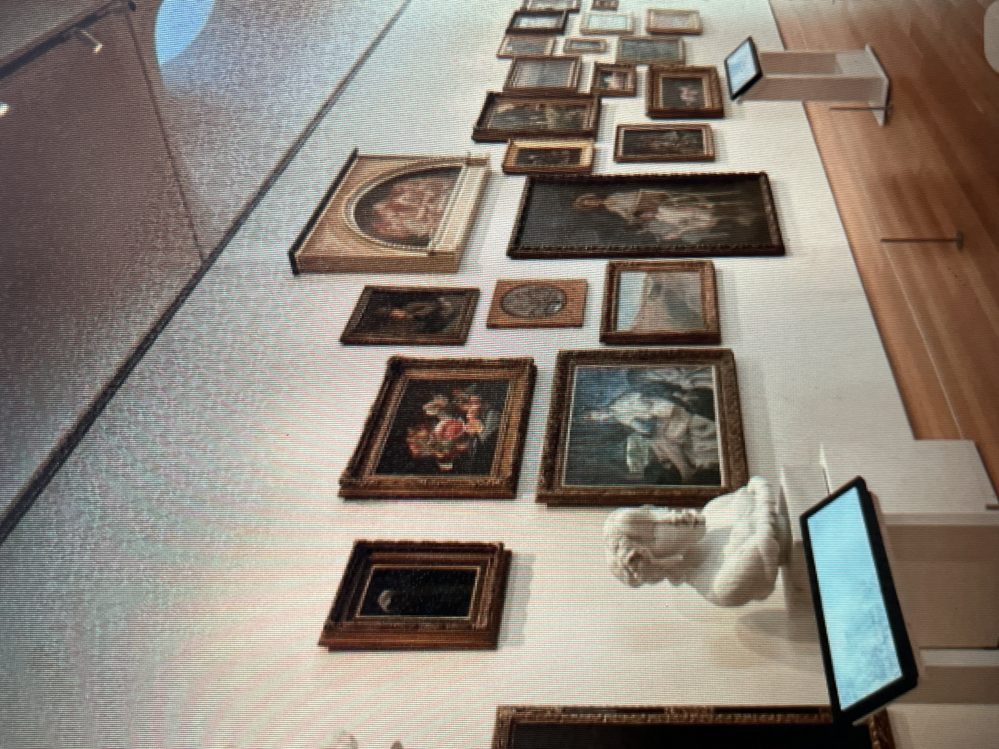 The image displays a museum exhibit with numerous framed artworks and a classical bust displayed on a pedestal. The artworks are hung on a white wall and vary in size and subject matter, including what appear to be still lifes and portraits. A modern digital display stand is positioned in the foreground, with a screen showing a bright blue image. The floor is made of polished wood. The image appears to be taken in an indoor setting, possibly a gallery, with artificial lighting. The location in Sejahtera, Indonesia is not specified within the visual information provided.