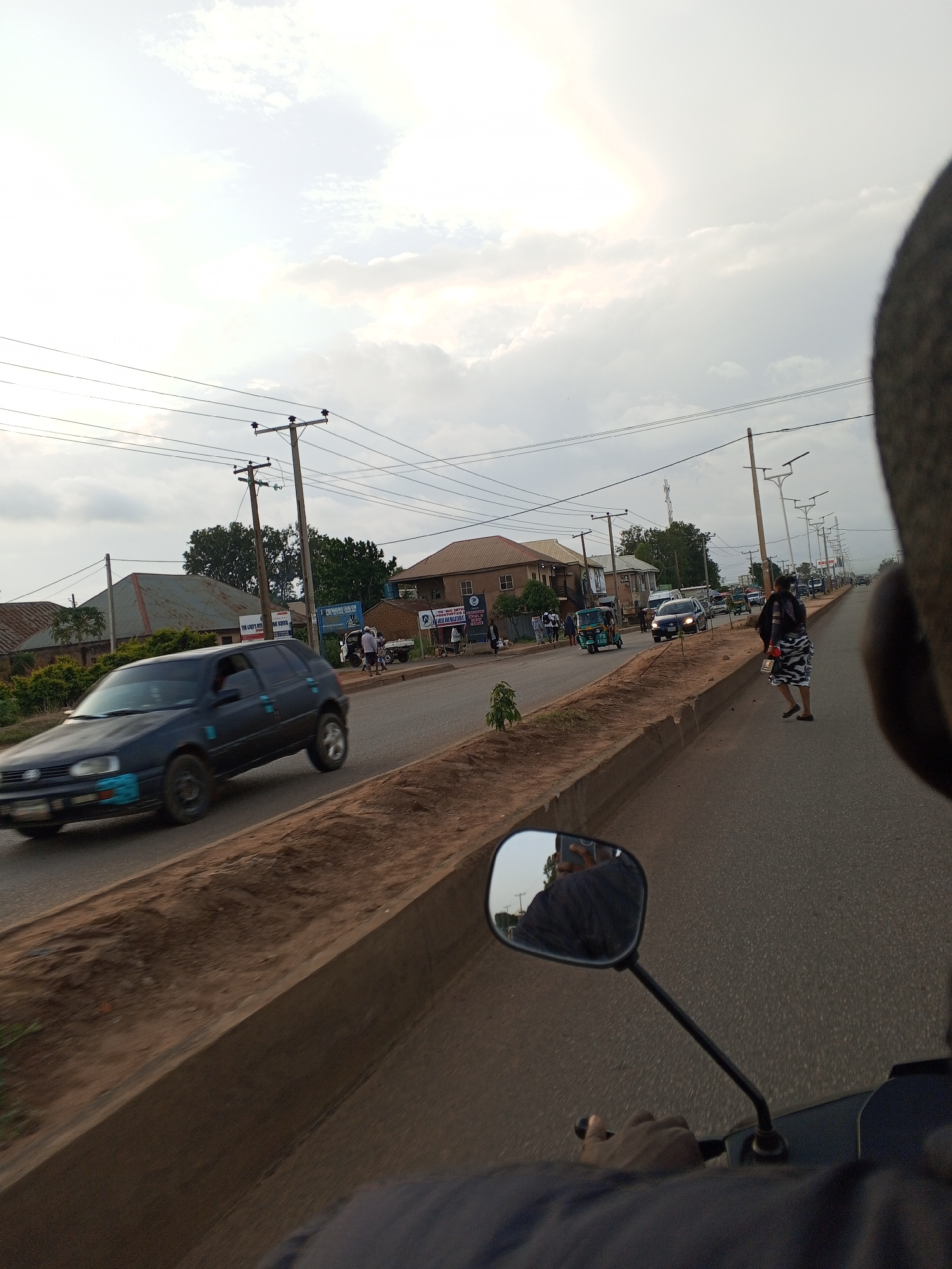 This media file shows a street scene in Bukuru, Nigeria, likely in the late afternoon given the soft, diffused light and long shadows. The weather appears overcast, with a bright sky peeking through the clouds. The setting is a paved road lined with a raised concrete median planted with some vegetation. On either side of the road, there are buildings, including houses and what appear to be shops or businesses, as well as utility poles with power lines stretching across the scene.
Several vehicles are visible, including a dark-colored hatchback car in motion on the left, a white truck, and a three-wheeled auto-rickshaw (keke napep) further down the road. People are also present: one person is walking on the sidewalk to the right, dressed in a patterned skirt and top, and others are seen near the buildings in the background. The perspective is from a vehicle, possibly a motorcycle, as indicated by the presence of a rearview mirror reflecting the rider and the road ahead.
Some text is visible on signs attached to the buildings. One sign appears to be for a school, with the words "FOR LEAPING" and "PRIMARY SCHOOL" partially legible. Another sign is for a business, with the letters "JN" and "UMIC ORTH" visible. The overall atmosphere suggests a typical day in a bustling Nigerian town.