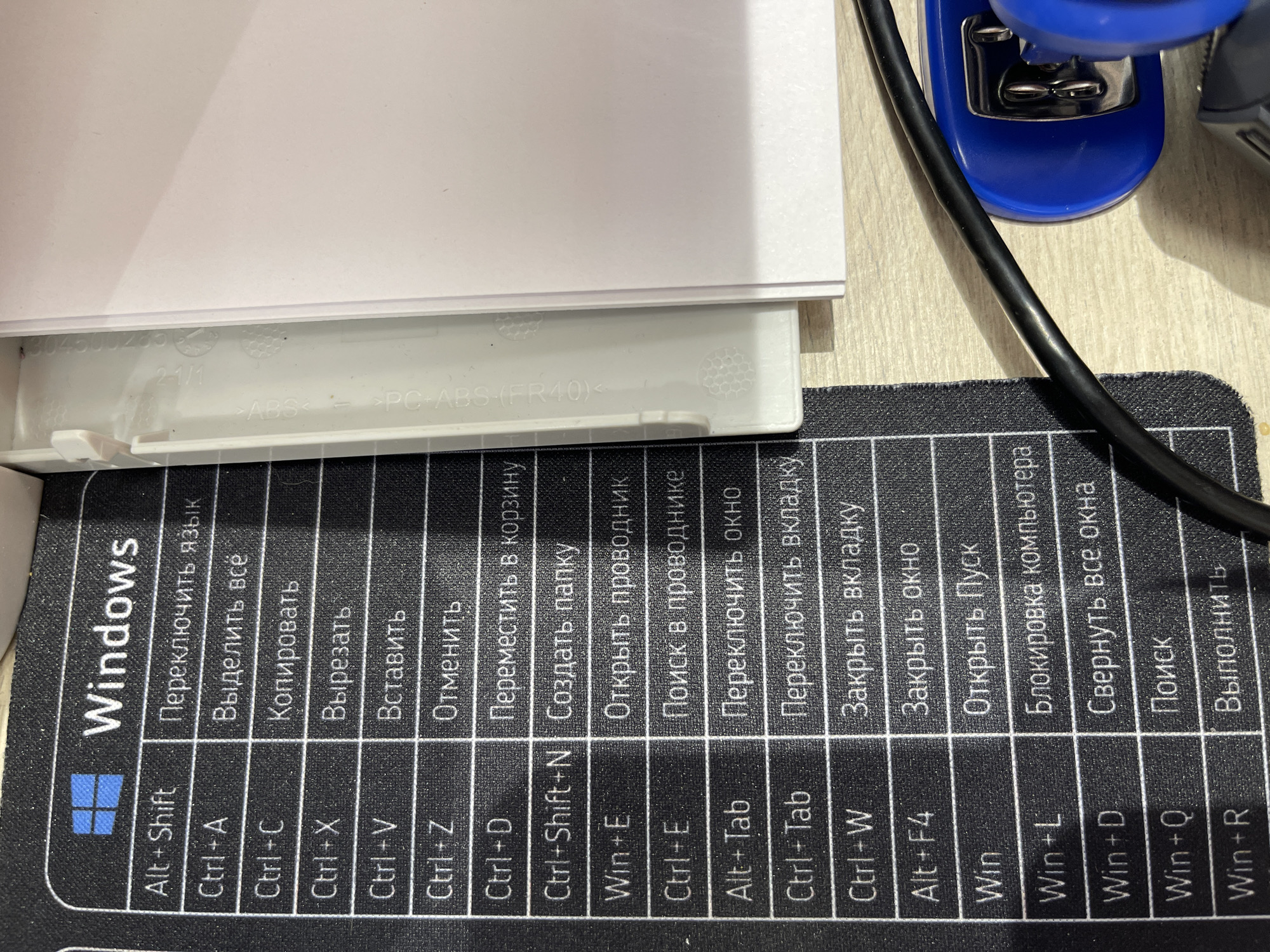 The image shows a close-up of a black mat with a white grid pattern and text. The text is in Russian and lists Windows keyboard shortcuts. The mat is placed on a light-colored wooden surface. To the right of the mat is a white object, possibly part of a device or furniture, with some text embossed on its surface. A black cable runs diagonally across the lower right portion of the image, leading to a blue object that appears to be an electronic device or accessory. The lighting suggests an indoor setting. There are no people visible in the image. The context provided about Moscow, Russia, is not visually verifiable from the image itself.