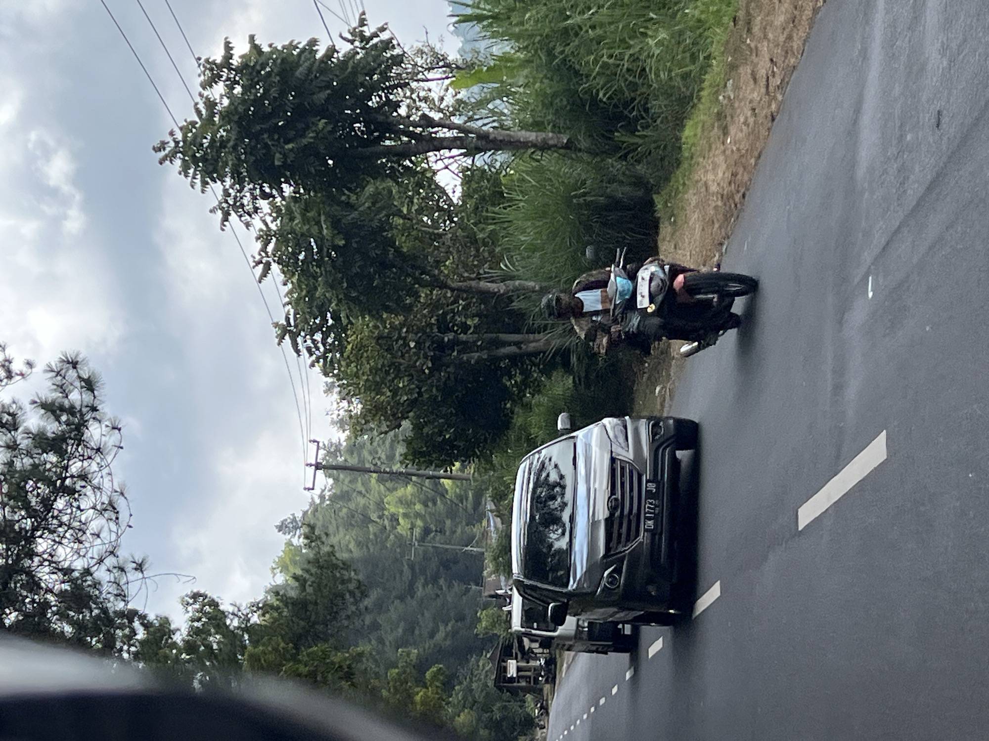 A grey SUV with license plate "DK 1773 JO" is on the left side of a two-lane asphalt road. The SUV is followed by a smaller white vehicle. On the right side of the road, a person wearing a helmet and a patterned jacket is riding a red and blue motorcycle. The road is surrounded by lush green vegetation and trees, with a cloudy sky above. The scene appears to be in a rural or mountainous area, consistent with the landscape around Kintamani, Indonesia.
