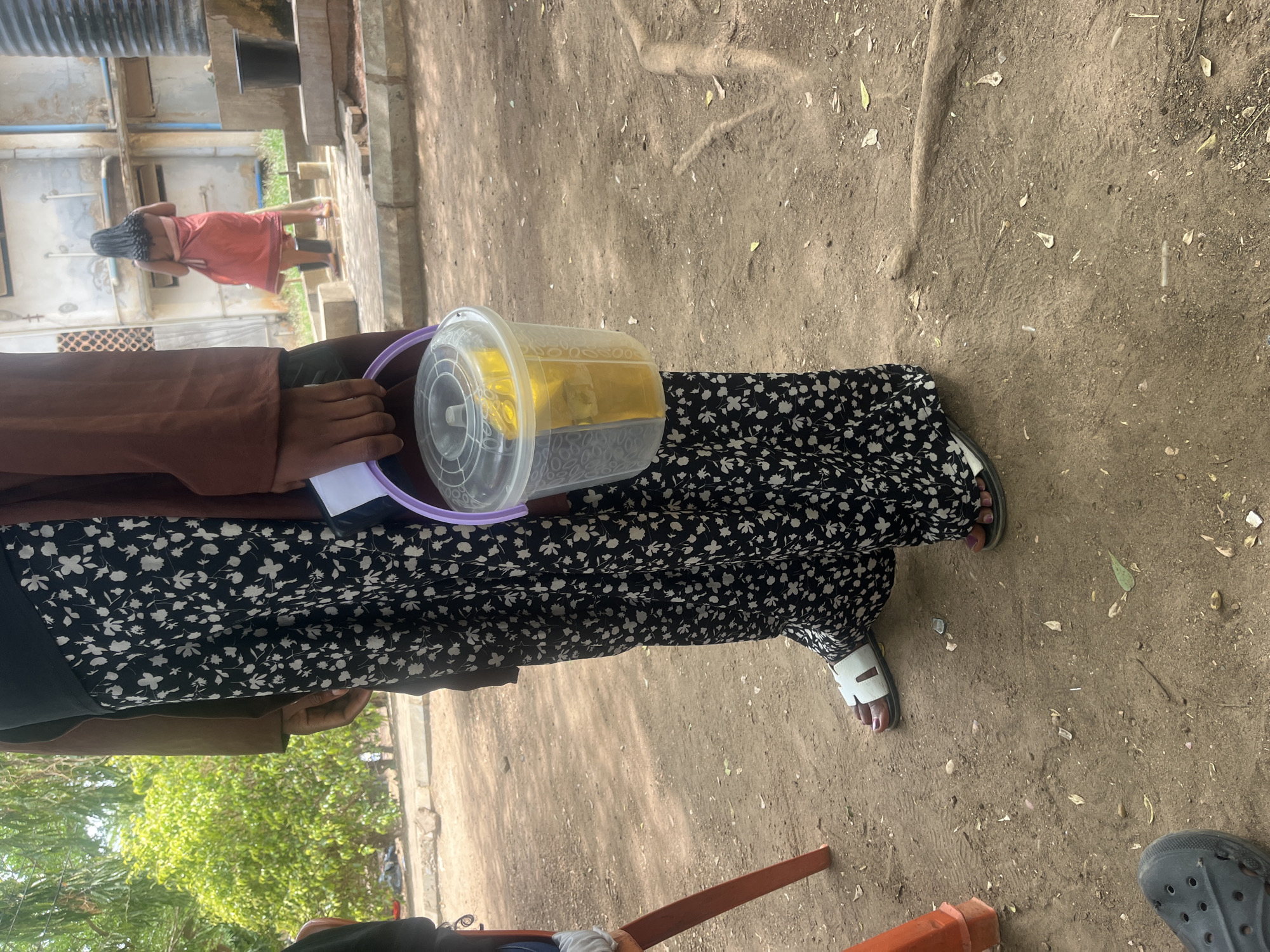 A ground-level shot of a person from the waist down, standing outdoors, likely in Unguwan Sanusi, Nigeria. The person is wearing a long black and white floral dress and sandals. They hold a clear plastic container with a purple handle, filled with a yellow substance, possibly food or liquid. The ground is dirt, and there are leaves and small debris scattered around. Another person is visible in the background near a building and is wearing a pink cloth. There's also a view of a green tree.