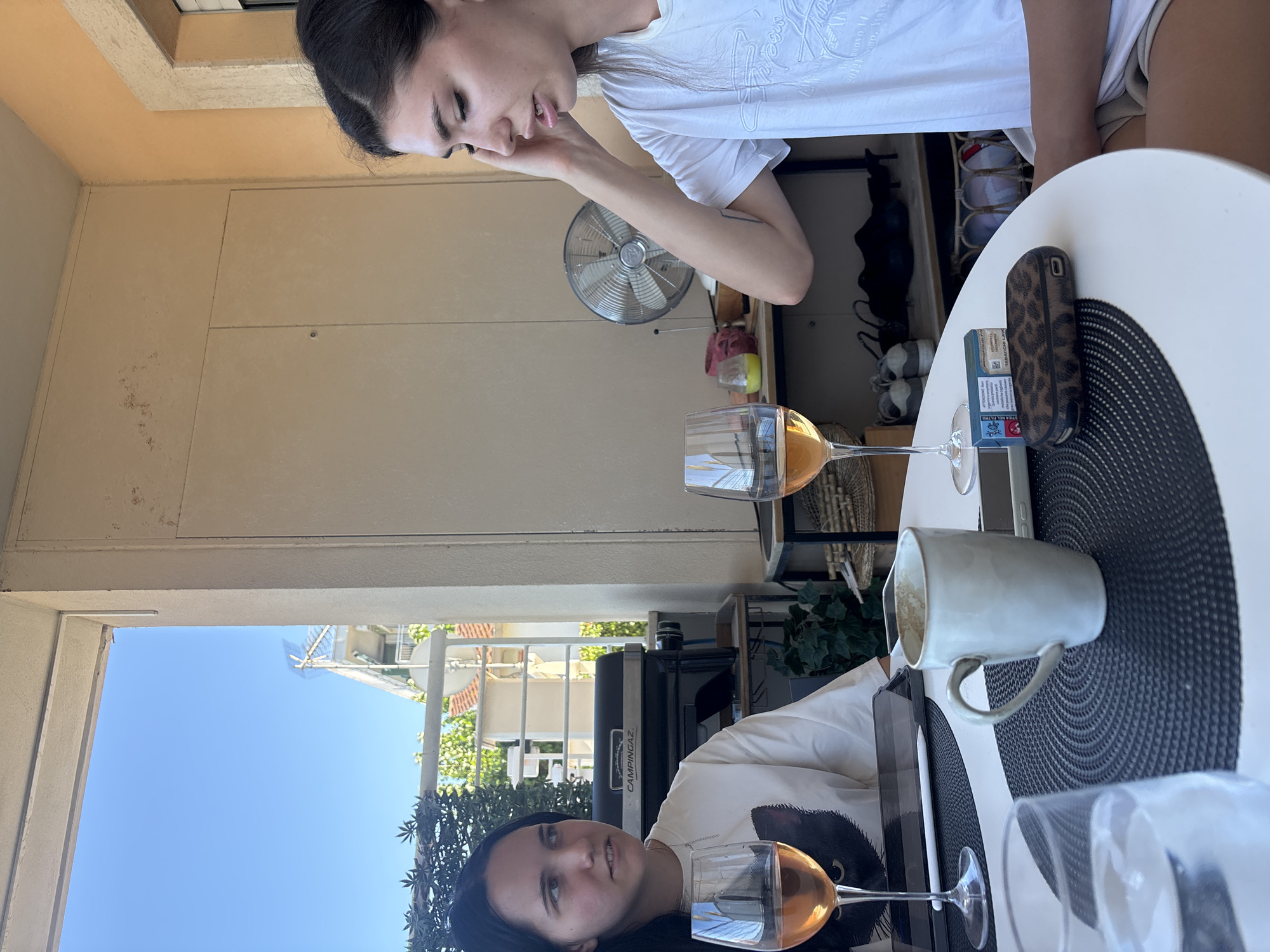Two young women are sitting at a round table on a balcony with a view of buildings and the sky. The woman on the left has dark hair and is wearing a white t-shirt with a black cat graphic. She is looking to her left. The woman on the right has dark hair pulled back and is wearing a white t-shirt. She is touching her forehead with her right hand and looking down.
On the table, there are two wine glasses filled with amber-colored liquid, a white mug, a tablet device, a pack of cigarettes, and a leopard-print case. In the background, there is a grill, a fan, and shelves with shoes and other items. The balcony is furnished with a white railing and has a beige-colored wall behind the woman on the right. The setting appears to be a residential building in Cupra Marittima, Italy, as indicated by the architecture and the view of residential buildings. The lighting suggests it is daytime.