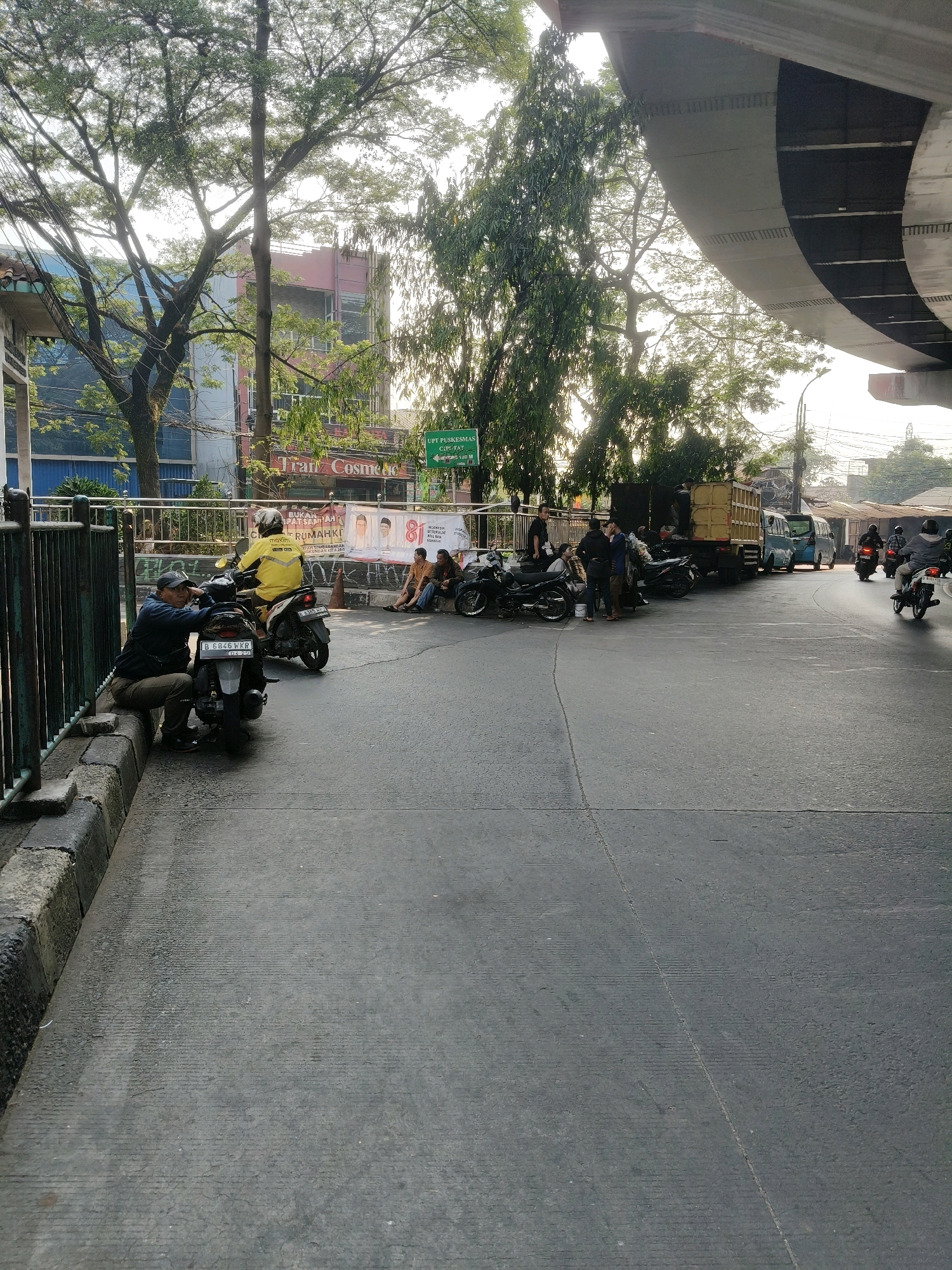 The image depicts a street scene in South Tangerang, Indonesia, under a highway overpass. In the foreground, a man wearing a dark jacket and cap is sitting on a concrete barrier next to a parked motorcycle with license plate B 6846 WXR. Another motorcycle, ridden by a person in a yellow jacket, is also parked nearby. A group of people are seated and standing further down the street, near parked motorcycles and a large yellow truck. Trees with dense foliage are visible throughout the scene, with a pink and blue building in the background. A green directional sign with Indonesian text is also present. The sky is bright, suggesting it is daytime. The overall environment appears to be an urban street with significant traffic infrastructure.