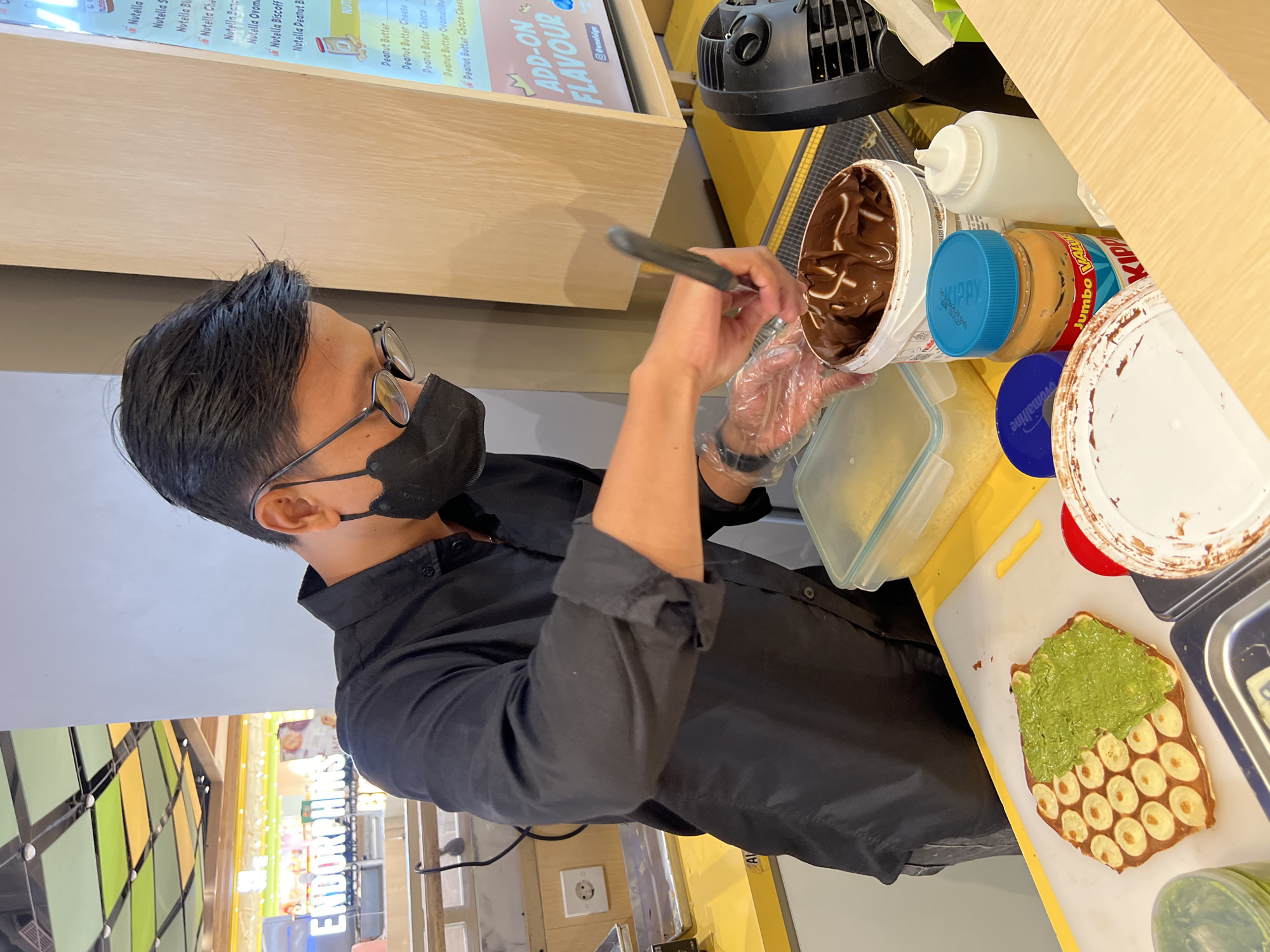 A male food vendor wearing a black shirt, black face mask, and glasses is preparing a bubble waffle with a green spread and banana slices on a white cutting board. He is holding a spatula with his right hand, which is gloved in clear plastic, and is scooping chocolate spread from a white bucket. In the background, a menu board displays various dessert options, including "Nutella" and "Peanut Butter" based items, and an "ADD-ON FLAVOUR" section. To the left, a sign reads "ENDORPHINS". A small fan is visible on the right. The scene appears to be inside a food stall or shop in Denpasar, Indonesia, based on the context of the "ENDORPHINS" sign, which is a known establishment in Bali. The overall environment is brightly lit, suggesting an indoor commercial setting.