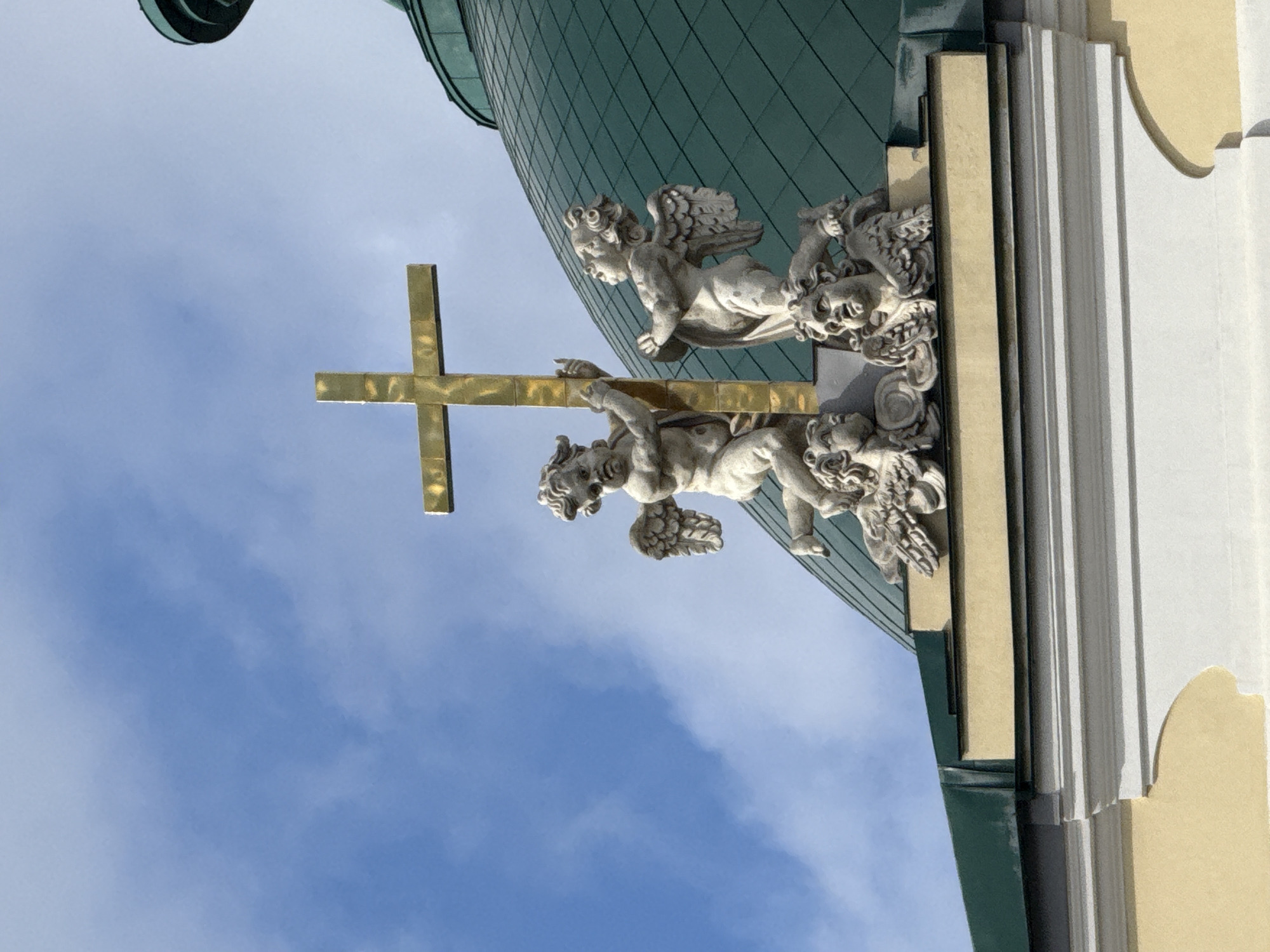 The image shows a golden cross held by two stone cherubs adorning a building, possibly a church or religious structure. The building has a green dome and decorative architectural elements, including moldings and light-colored walls. The background is a blue sky with some clouds. The architectural style appears to be European, potentially Baroque or Rococo. Based on the style and architecture, it could be located in St. Petersburg, Russia.