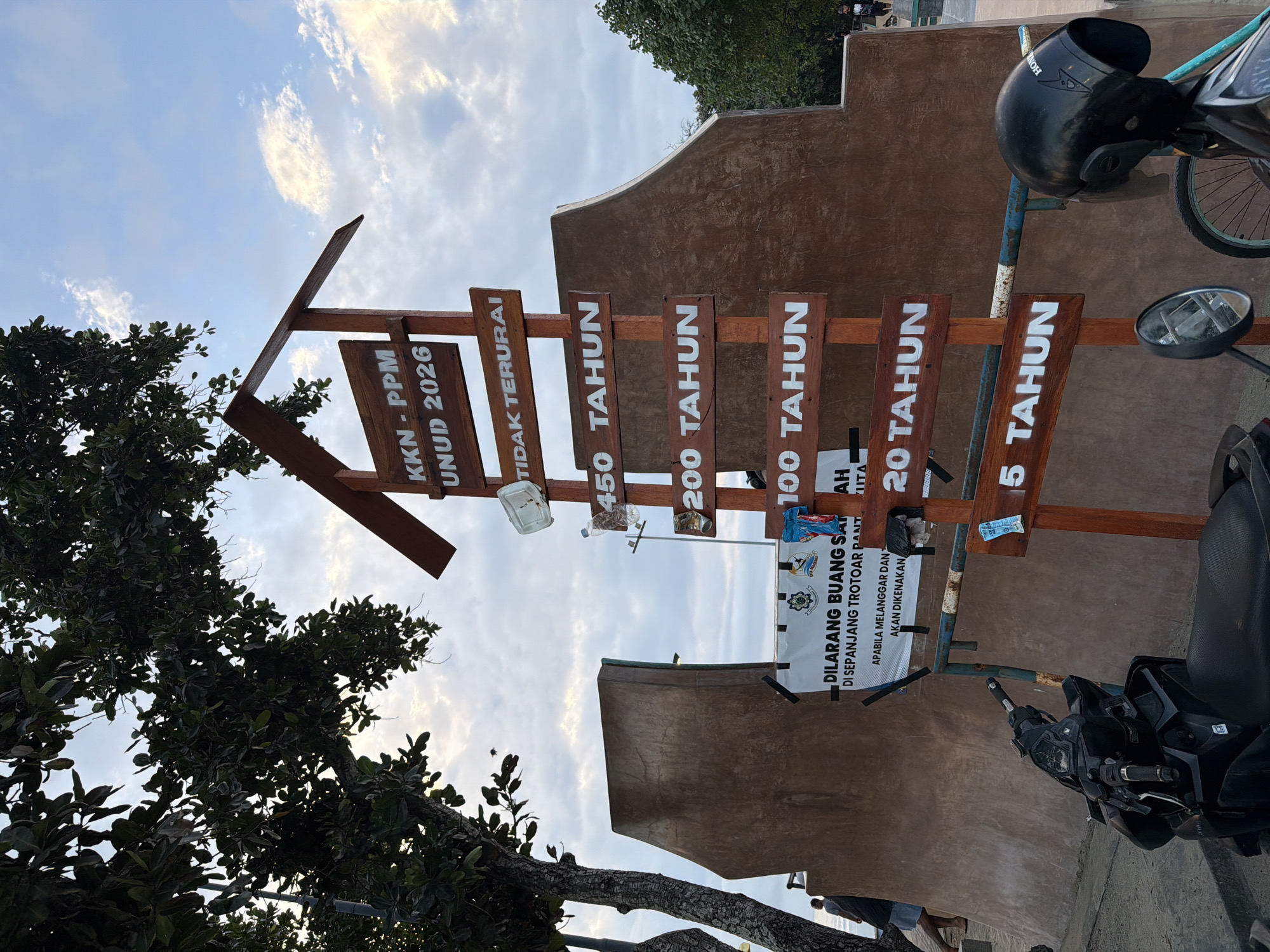 The image shows a sign in Kuta, Indonesia, with an arrow shape pointing upwards. The sign consists of several wooden planks, each labeled with a number of years: "KKN-PPM UNUD 2026," "TIDAK TERURAI," "450 TAHUN," "200 TAHUN," "100 TAHUN," "20 TAHUN," and "5 TAHUN." Below these planks, a poster reads "DILARANG BUANG SARAH DI SEPANJANG TROTOAR PANT," indicating a prohibition against littering. A motorcycle is parked next to the sign. The background features a brown wall and glimpses of trees and sky.