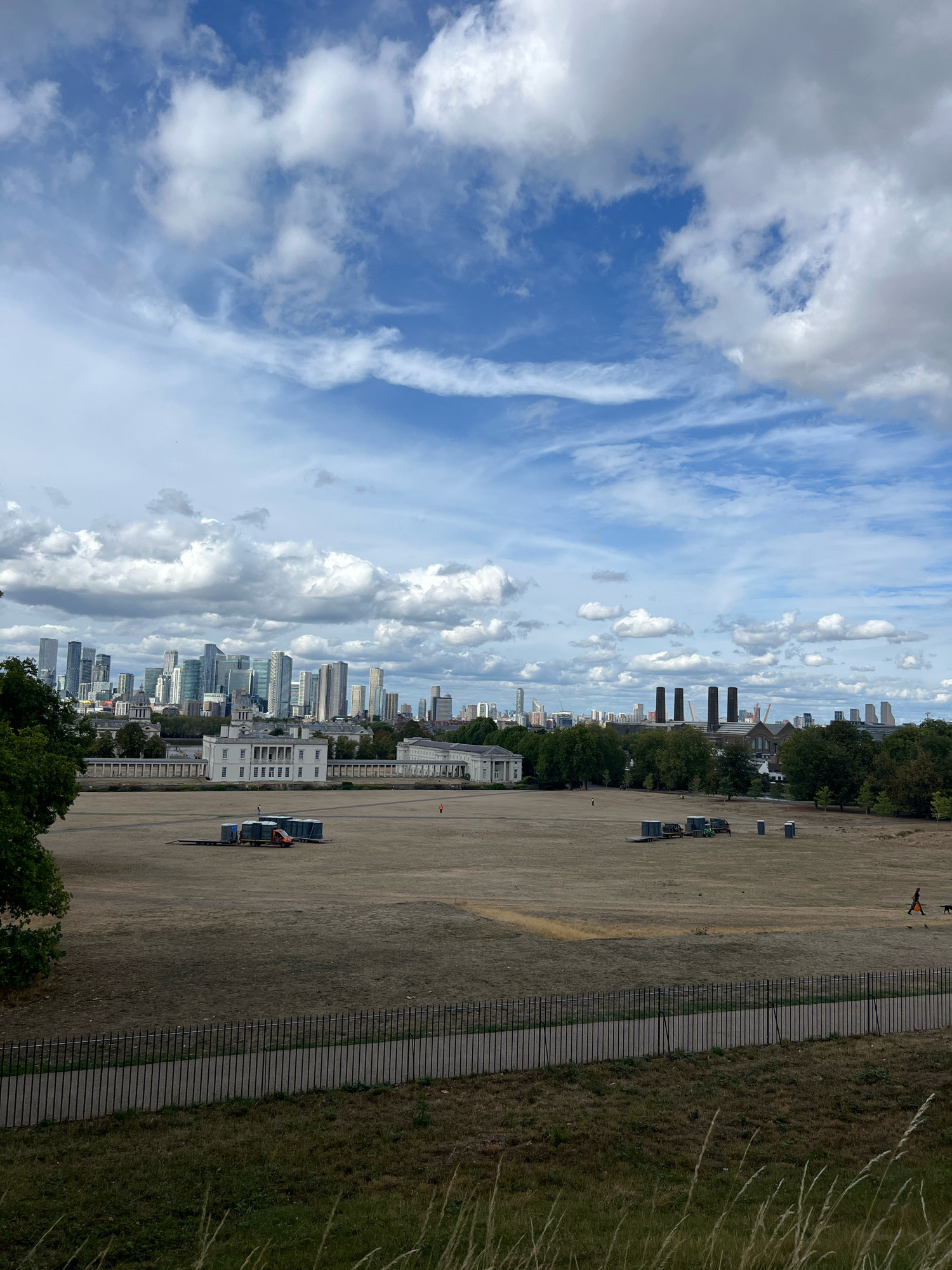The image depicts a panoramic view of London from an elevated vantage point, likely Greenwich Park, overlooking the Canary Wharf skyline and the River Thames. The foreground features a wide expanse of dry, brown grass, indicative of a dry summer. A black metal fence runs horizontally across the lower portion of the image, separating the grassy hill from a paved walkway. Beyond the field, a long, white classical building with a colonnade stretches across the middle ground, possibly part of the Royal Observatory or the National Maritime Museum. In front of this building, several portable toilets and a small truck are situated on the dry grass. Further into the distance, the dense urban landscape of Canary Wharf rises, characterized by numerous modern skyscrapers. To the right of the frame, a person is walking a dog on a leash, and another figure in an orange vest is visible further away. The sky is a mix of blue and scattered white clouds.