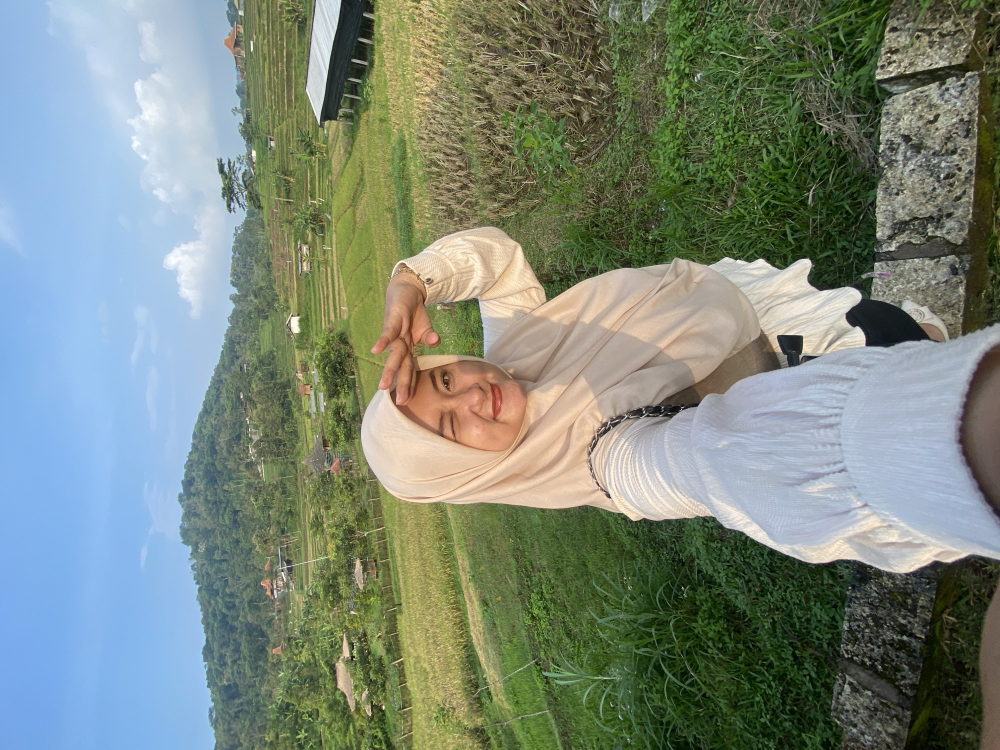 A young woman in a hijab poses for a selfie. She is wearing a beige hijab and a long-sleeved, white, crinkled shirt. She is standing in front of rice terraces on a hillside near Penanggungan, Indonesia. A mountain is visible in the background. The sky is blue.