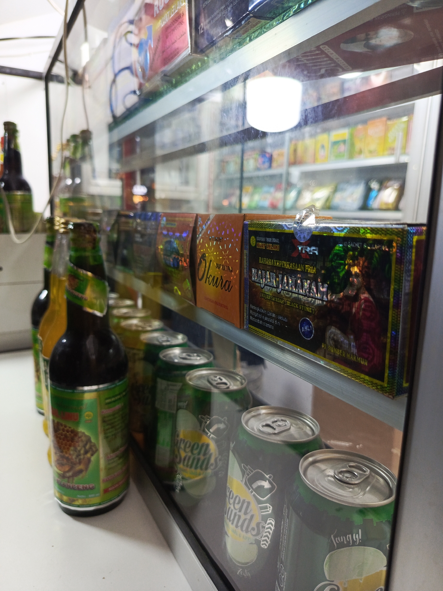 This image depicts a close-up view of a display shelf inside a convenience store, likely a minimarket, located in Tangerang, Indonesia. The shelf is made of glass and metal, showcasing various packaged goods and beverages. On the left, several green-brown glass bottles are visible, one in the foreground with a green and gold label featuring an illustration of a honeycomb and text. Adjacent to these are several cans of "Green Sands" soda, characterized by their green color and prominent branding with cartoonish designs. In the background, behind the glass, a variety of colorful packaged items are arranged on shelves, with some appearing to be food products or health supplements. The lighting in the store appears to be artificial, with a bright white light source visible in the upper portion of the frame. The overall environment suggests a commercial setting designed for retail sales.