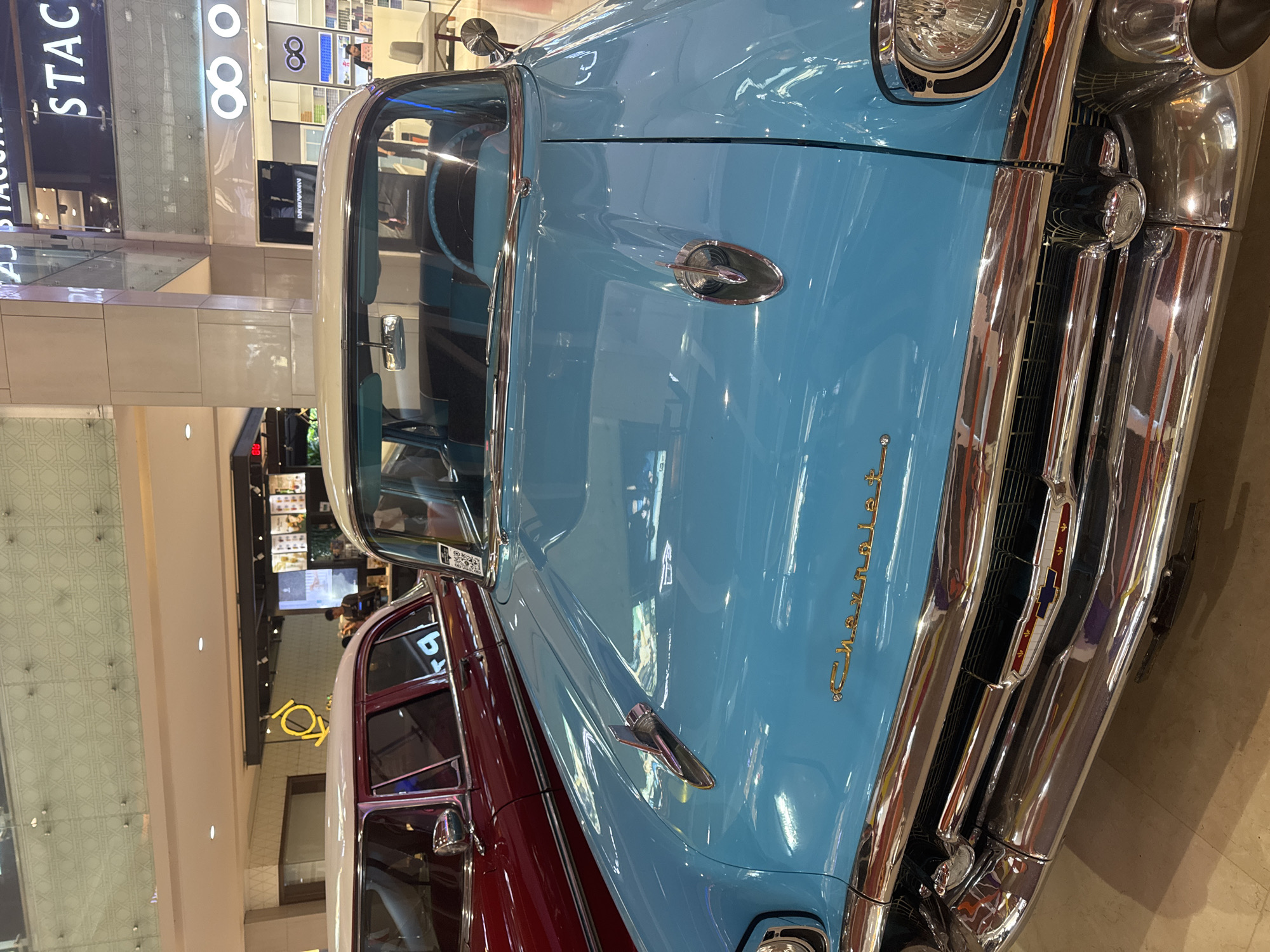 The image shows a close-up, front view of a classic Chevrolet car, parked indoors. The car has a two-tone color scheme, with a light blue hood and a maroon body. Chrome detailing is prominent on the grill, bumper, and other parts of the car. The car is located inside a building, likely a shopping mall, given the presence of store signs and the general architectural design of the space. The location is a city named Unknown, Unknown.