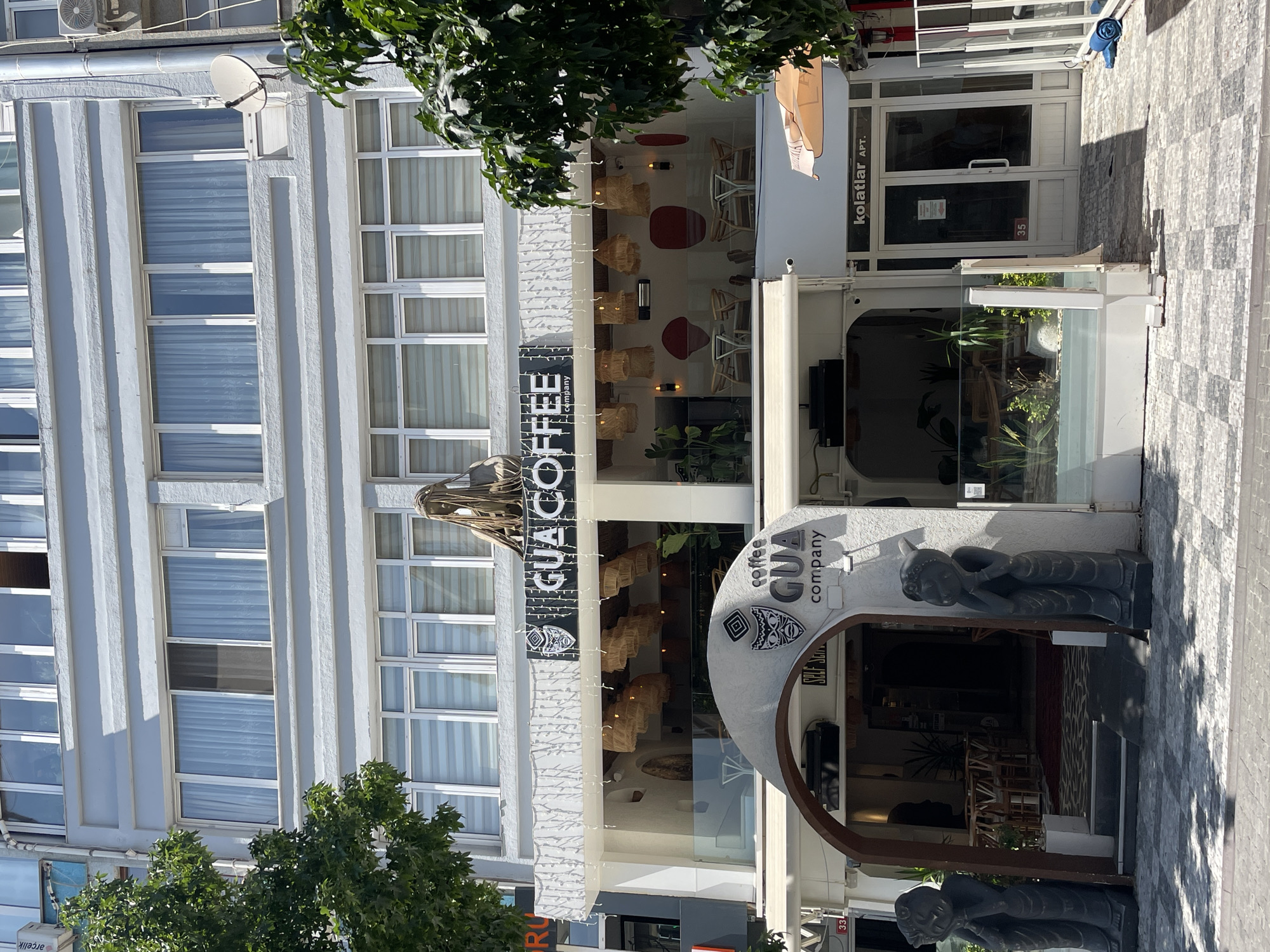 A bright, sunny day illuminates a multi-story building with a light grey facade. The building features numerous windows with white frames, some covered by sheer white curtains and others with roller blinds. The ground floor is occupied by a business with a white awning and a prominent sign reading "GUA COFFEE company" with a stylized tribal mask logo. This entrance is framed by an arched opening and flanked by two dark grey, seated human-like statues. Beyond the arch, the interior of the establishment is visible, showcasing tables, chairs, and lush green plants. To the right of the coffee shop entrance, a white door with a sign that reads "kolatlar APT." suggests residential units above or adjacent. The street in front is paved with a checkered pattern of light and dark grey tiles. Trees with vibrant green foliage frame the scene on the left and right, casting dappled shadows on the pavement. The overall impression is of a bustling urban commercial area.