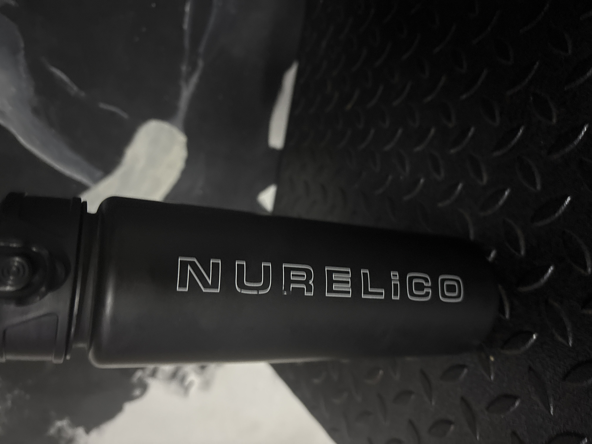 A black reusable water bottle with "NURELICO" inscribed vertically in white capital letters is positioned in the foreground. The bottle is cylindrical with a textured lid. It rests on a black, textured surface, possibly a gym floor or industrial platform, with a raised diamond pattern. In the background, a blurred abstract image, possibly a painting or mural, is visible against a dark wall. The overall lighting is dim, with some highlights on the bottle and the background artwork. The image does not contain any identifiable location information or signs indicating it is in Orsk, Russia.