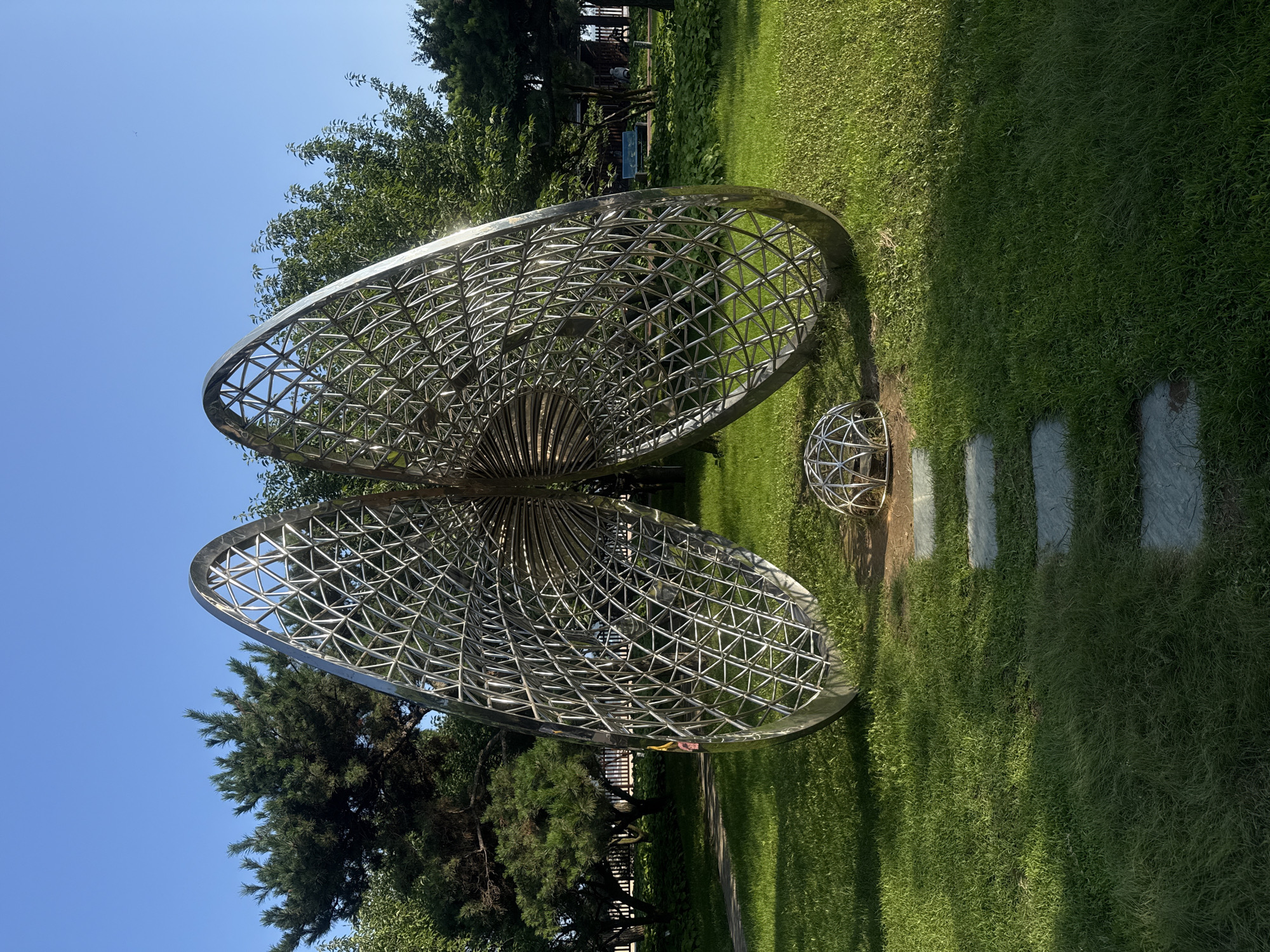 A large, silver, butterfly-shaped sculpture made of geometric metal latticework stands in a grassy park on a sunny day. Two large, curved panels resembling butterfly wings rise from a central base, connected by a series of concentric circular elements. At the base of the sculpture, a smaller, spherical metal lattice structure rests on the ground. A path of flat, square stones leads towards the sculpture from the foreground. The park is lush with green grass and trees, including pine trees, and a clear blue sky is visible above. In the background, a fence and some distant figures suggest a public area. The sculpture is located in Beidaihe District, Qinhuangdao City, Hebei Province, China.