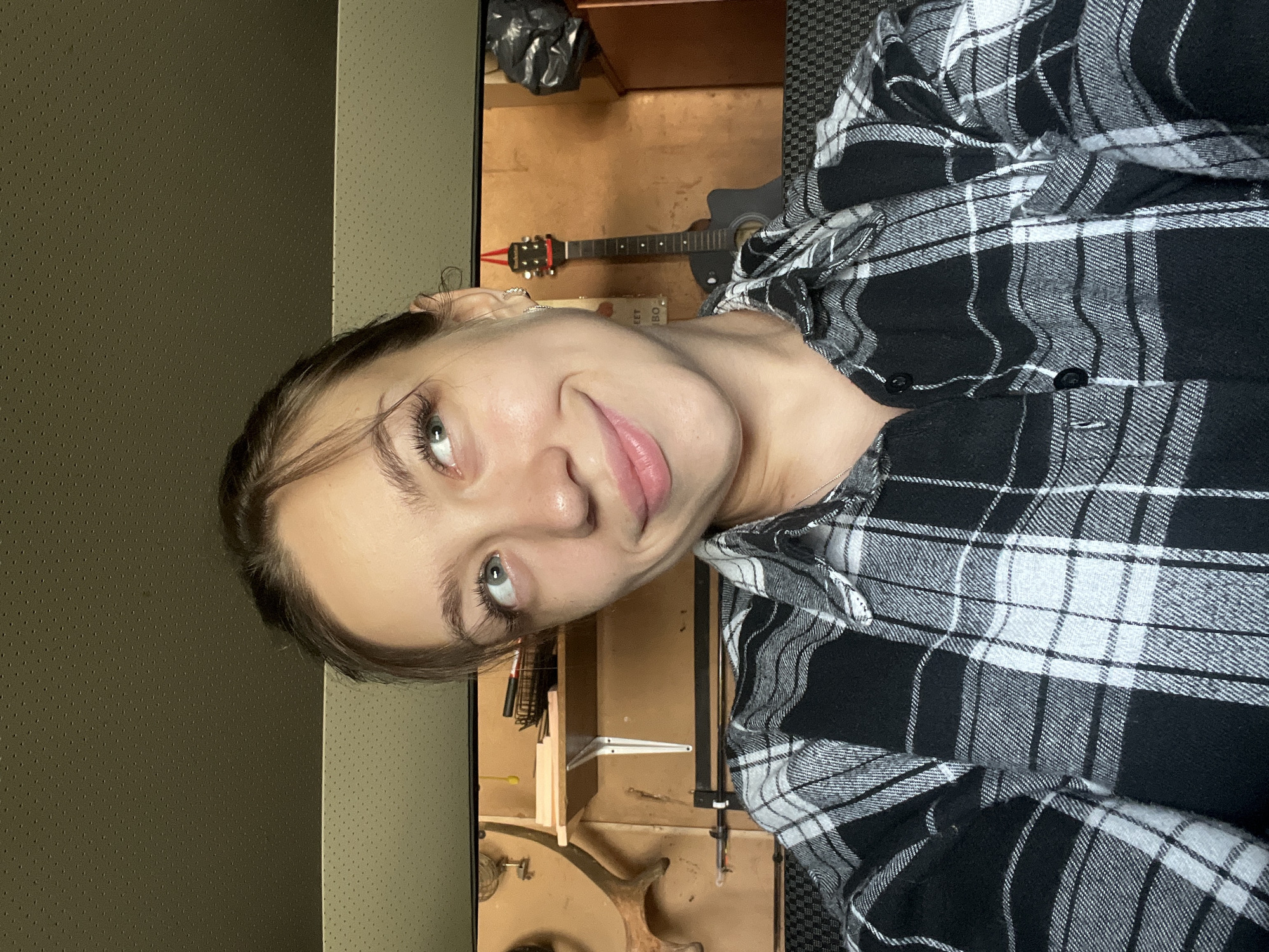 The image shows a young woman with light-colored eyes and dark hair pulled back, looking upwards and to her right with a slight smile. She is wearing a black and white plaid shirt. The background appears to be an indoor space with shelves containing various objects, including what looks like a deer antler and a guitar. The overall impression is a casual, informal setting. The location is specified as Kemul, Russia.