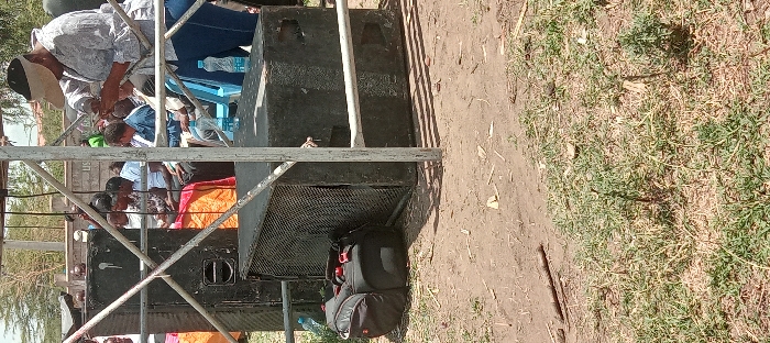 The image shows a scene in Kitengela, Kenya, at the coordinates provided. The scene is dominated by two large, black speaker cabinets. The cabinets are positioned behind a metal scaffolding structure. There is a black bag lying on the ground next to the speaker cabinets. Several people are seated in the background, partially obscured by the scaffolding. There is also a water bottle visible in the foreground. The ground is dirt, with patches of grass and some dried vegetation. There are trees visible in the background.