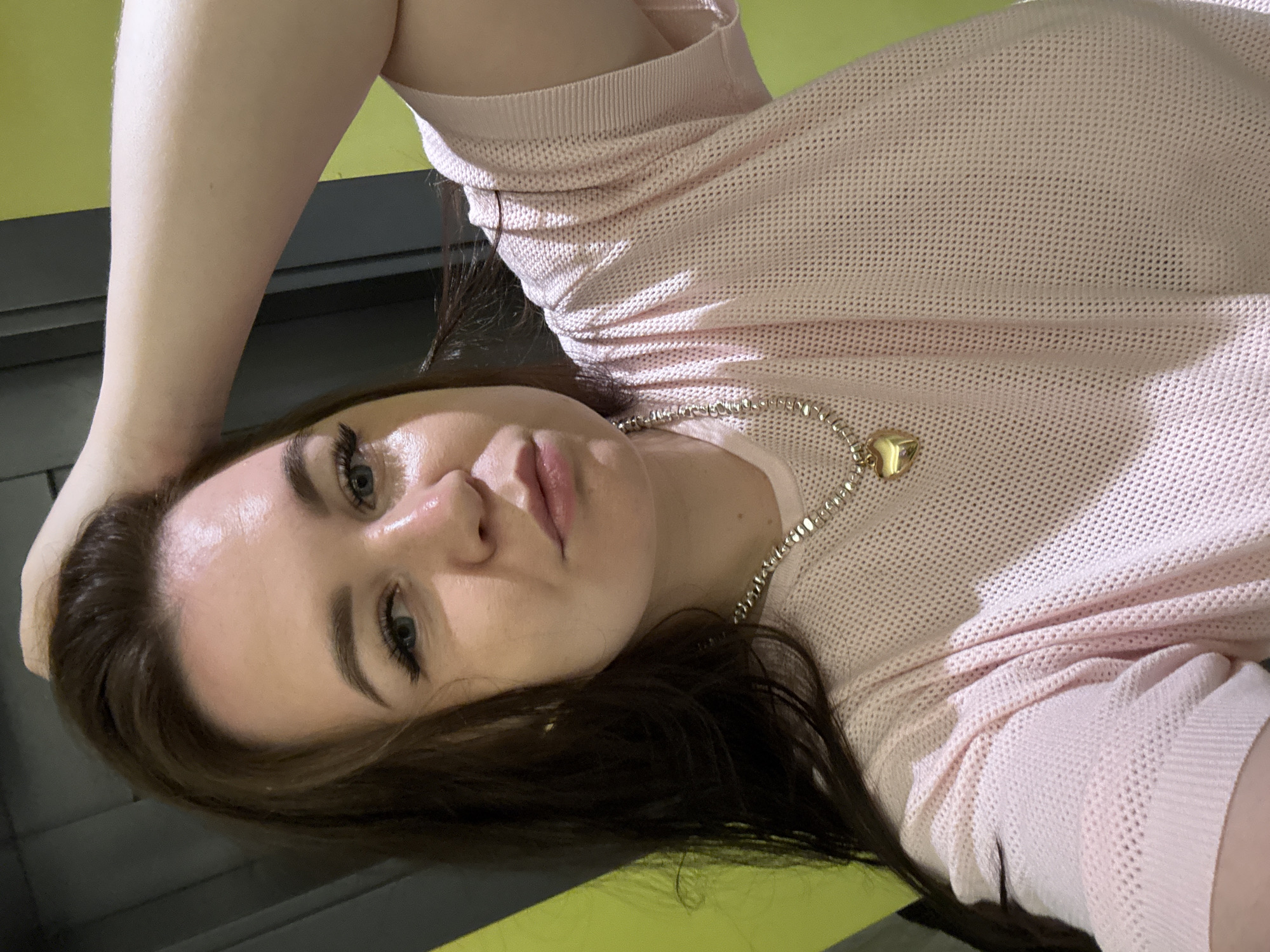 An overhead selfie shows a woman with long brown hair touching her head. She is wearing a light pink, mesh-like sweater and a silver necklace with a gold heart pendant. The background is blurred, with a lime green wall on the left and a dark gray wall or door on the right. The scene appears to be indoors, possibly in a modern setting. There is no information available to place this image in Alanya, Turkey.