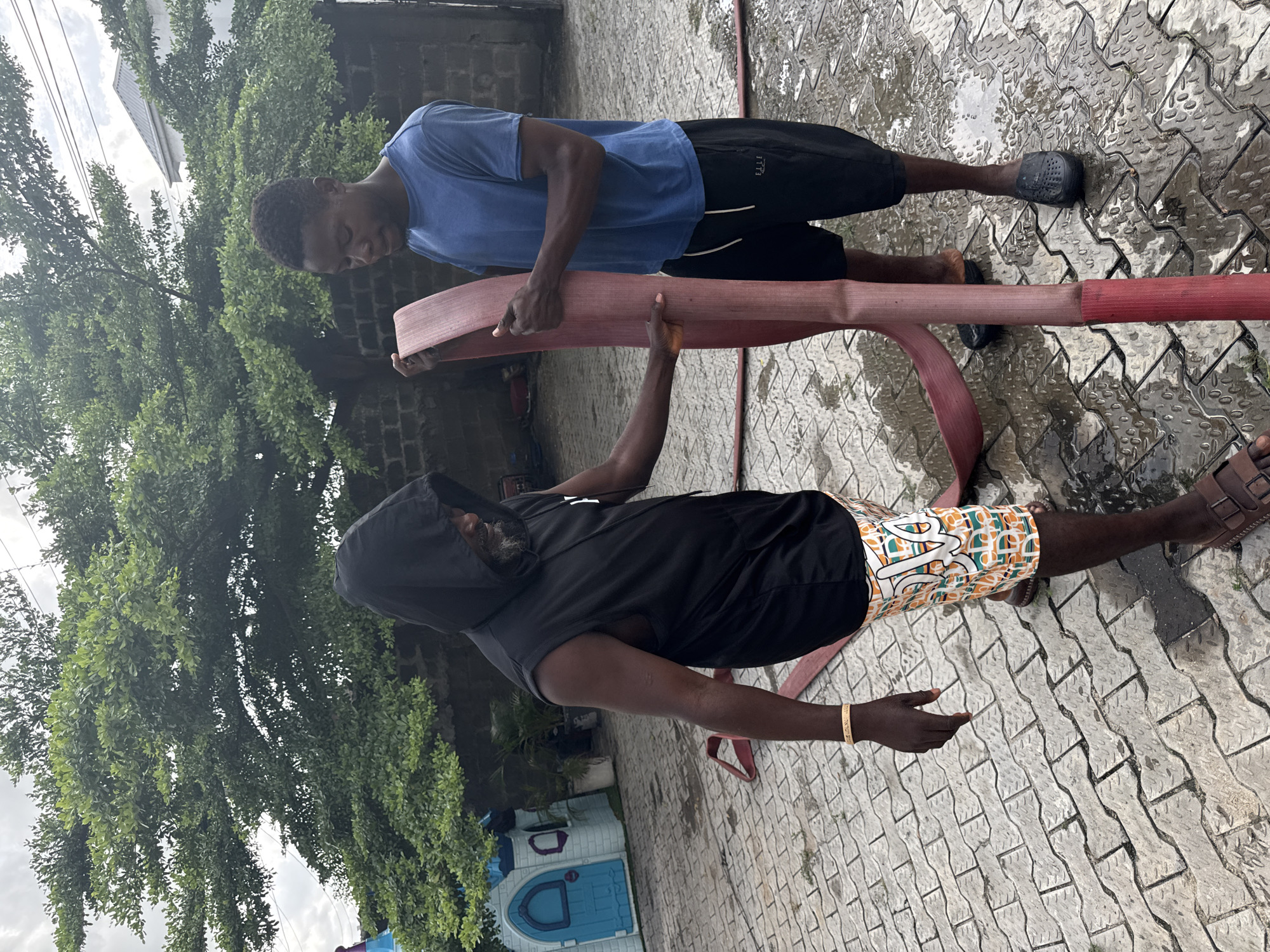 Two men stand on a paved surface outside, near a tree and a small blue-and-white building, likely in Ado, Nigeria. They are holding a flattened, red fire hose. One man wears a blue shirt and black shorts, while the other wears a black hooded top and patterned shorts. The ground is wet, possibly from recent use of the hose.