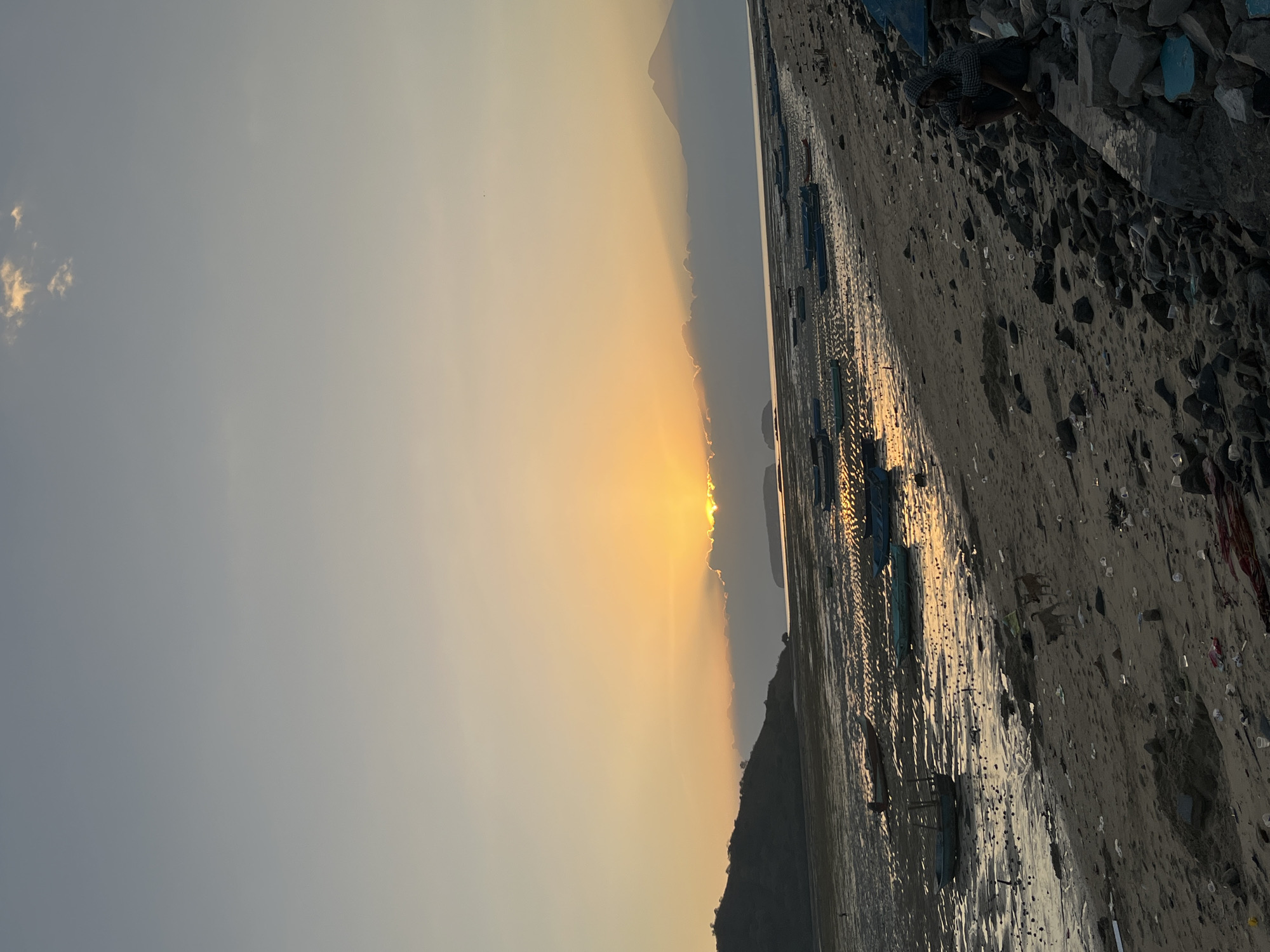 The image captures a sunset scene at the beach in Kertasari, Indonesia. The sun is low on the horizon, partially obscured by clouds, casting a warm golden light across the sky and reflecting on the wet sand and shallow water left by the receding tide. Numerous small boats are scattered across the intertidal zone, some resting on the sand and others partially submerged. The shoreline is littered with debris, including plastic bottles and other trash. In the foreground, on the right side of the image, a person wearing a hooded jacket sits on a pile of rocks, looking down at something in their hands. Further down the beach, two dogs are visible, one standing and one walking. In the background, a silhouette of a mountain is visible against the sunset sky, with a faint outline of a smaller island or landmass also present. The overall impression is one of a quiet, somewhat neglected beach during sunset.