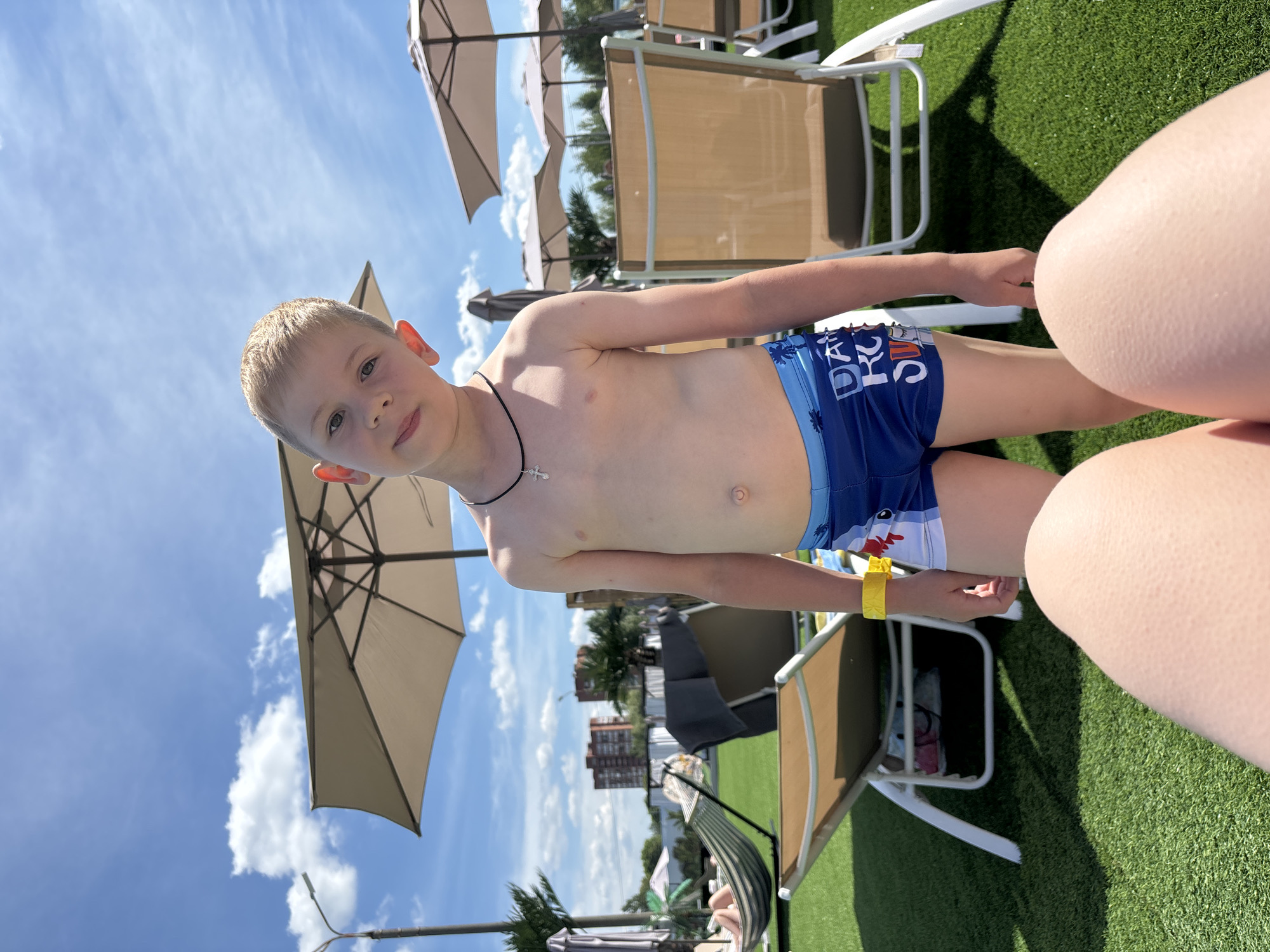 A young boy, appearing to be around 5-7 years old, is the central subject in this image. He has short, light-colored hair and is looking directly at the camera with a neutral expression. He is wearing blue swim trunks with a pattern of sharks and the text "DA... RU..." is visible on the trunks. He is also wearing a thin black necklace with a small cross pendant and a yellow wristband.
The boy is standing in an outdoor setting, likely a pool or resort area, as indicated by the artificial green turf, lounge chairs, and large umbrellas. In the background, buildings are visible, suggesting an urban or developed area. The sky is bright blue with scattered white clouds, and the lighting suggests it is daytime, likely sunny. The image is shot from a low angle, looking up at the boy. The perspective also suggests that the photographer is either lying down or very close to the ground. There is a blurred portion of another person's leg in the lower right corner of the frame.
