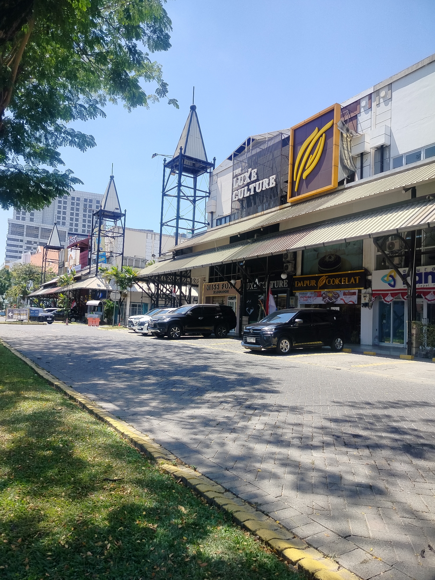 The image shows a sunny day in Surabaya, Indonesia, with a view of a street lined with shops and buildings. In the foreground, a paved road is visible, bordered by a grassy area with trees casting shadows. Several cars are parked along the road in front of the shops. The shops have awnings and signs, some of which are in Indonesian. One sign reads "DAPUR COKELAT" which translates to "Chocolate Kitchen". In the background, tall buildings suggest an urban environment. The sky is clear and blue.