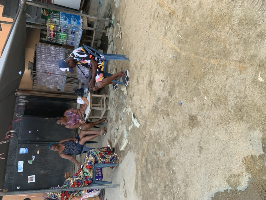 This image depicts an outdoor scene in Ikota, Nigeria, under bright daylight, showing four individuals gathered on an unpaved, sandy ground, likely in front of a small shop or residential area.
In the mid-ground, a man wearing a colorful patterned short-sleeved shirt, shorts, a baseball cap, and sandals is seated on a blue plastic chair. He leans forward, seemingly engaged with objects on a small, light-colored wooden table positioned in front of him. To his left, two women are also seated on blue plastic chairs. The woman closer to the man is dressed in a patterned top and shorts, looking towards him. Next to her, another woman in a dark top and shorts faces slightly away. Partially visible on the far left, a fourth woman wears a long, vibrantly patterned skirt and a dark top, seated on another plastic chair.
The background features a small, informal shop or stall with shelves displaying numerous bottles of water and other beverages. Dark metal gates or shutters are also visible, suggesting a closed or partially enclosed structure. The ground is a mix of sand and dirt, with some small debris scattered. The overall atmosphere appears casual and relaxed, indicative of a typical local gathering or informal commercial setting. No specific text on product labels or other signs is clearly legible.