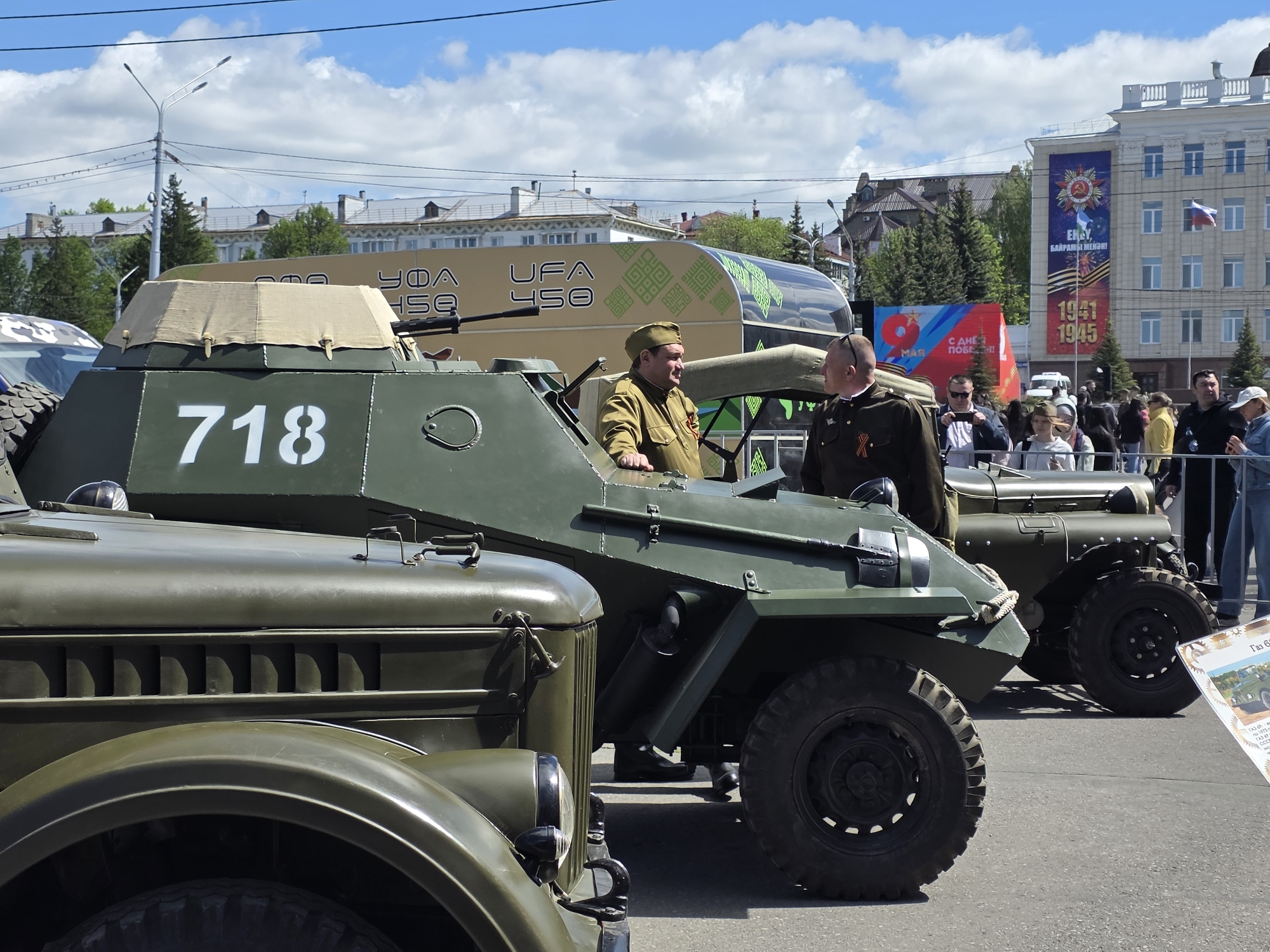 The image shows a street scene in Ufa, Russia, featuring several vintage military vehicles on display. The most prominent vehicle is a green armored car with the number "718" painted on its side. Two men in military-style uniforms are standing near the vehicle, possibly engaging in conversation. In the background, there is a building with banners commemorating Victory Day (May 9th) and celebrating the 450th anniversary of Ufa. Several people are visible in the background, likely spectators or attendees of the event. The sky is partly cloudy.