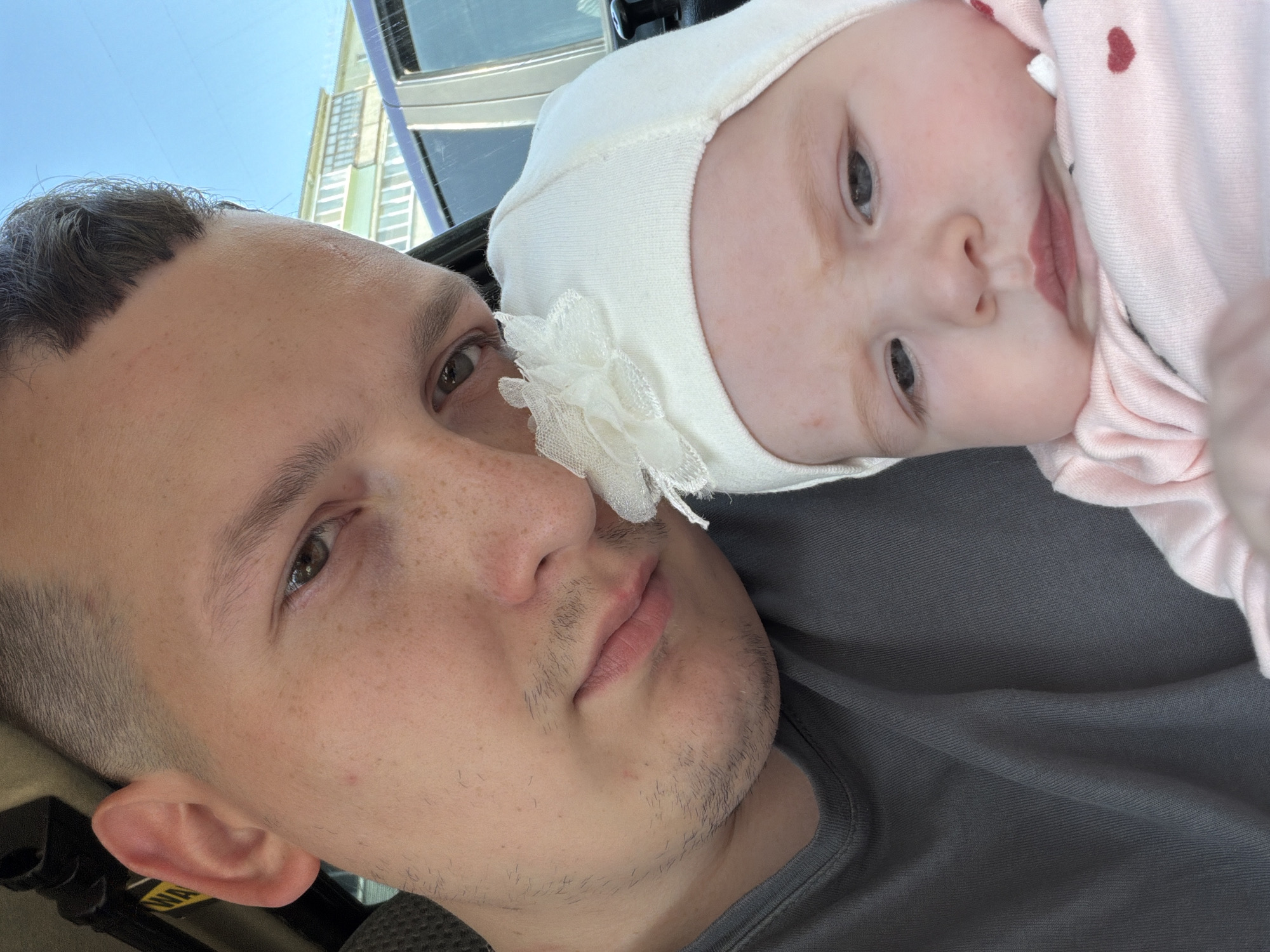 A close-up selfie shows a man and a baby in a car. The man, on the left, has short hair and is wearing a grey t-shirt. He is looking at the camera with a serious expression. The baby, on the right, is wearing a white hat with a flower on it and a pink outfit. The baby is looking away from the camera with a pensive expression. In the background, through the car window, a light-colored building with balconies and a blue sky are visible. The scene appears to be in the city of Noviy, Russia.