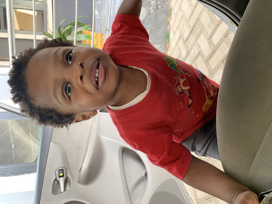 A child in a red shirt with cartoon characters is in a car. The child is standing with their arm extended, leaning on the car door. The car door is white and has a speaker grill. The car seat is olive green. The background shows light-colored brick flooring and a plant. The child is looking directly at the camera with a slight smile, and their head is tilted. The child has dark hair and eyes.
