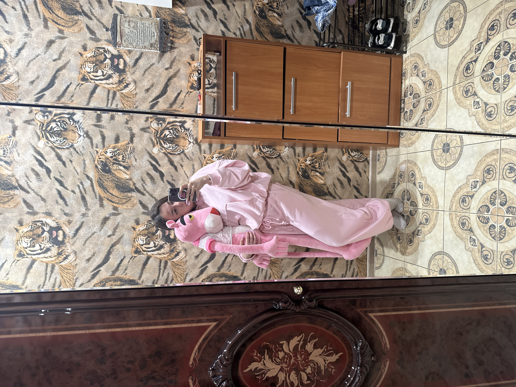 A person is lying on a tiger-print rug, holding a large pink stuffed panther. They are wearing a pink tracksuit and holding a smartphone. The scene appears to be indoors, possibly a bedroom or living room, with a wooden cabinet and tiled floor visible. The lighting suggests daytime. The reflection in what appears to be a mirrored surface shows the person and their surroundings. The backdrop is a tiger print wallpaper. The person's expression is neutral. There is no visible text. The location is Magnitogorsk, Russia.