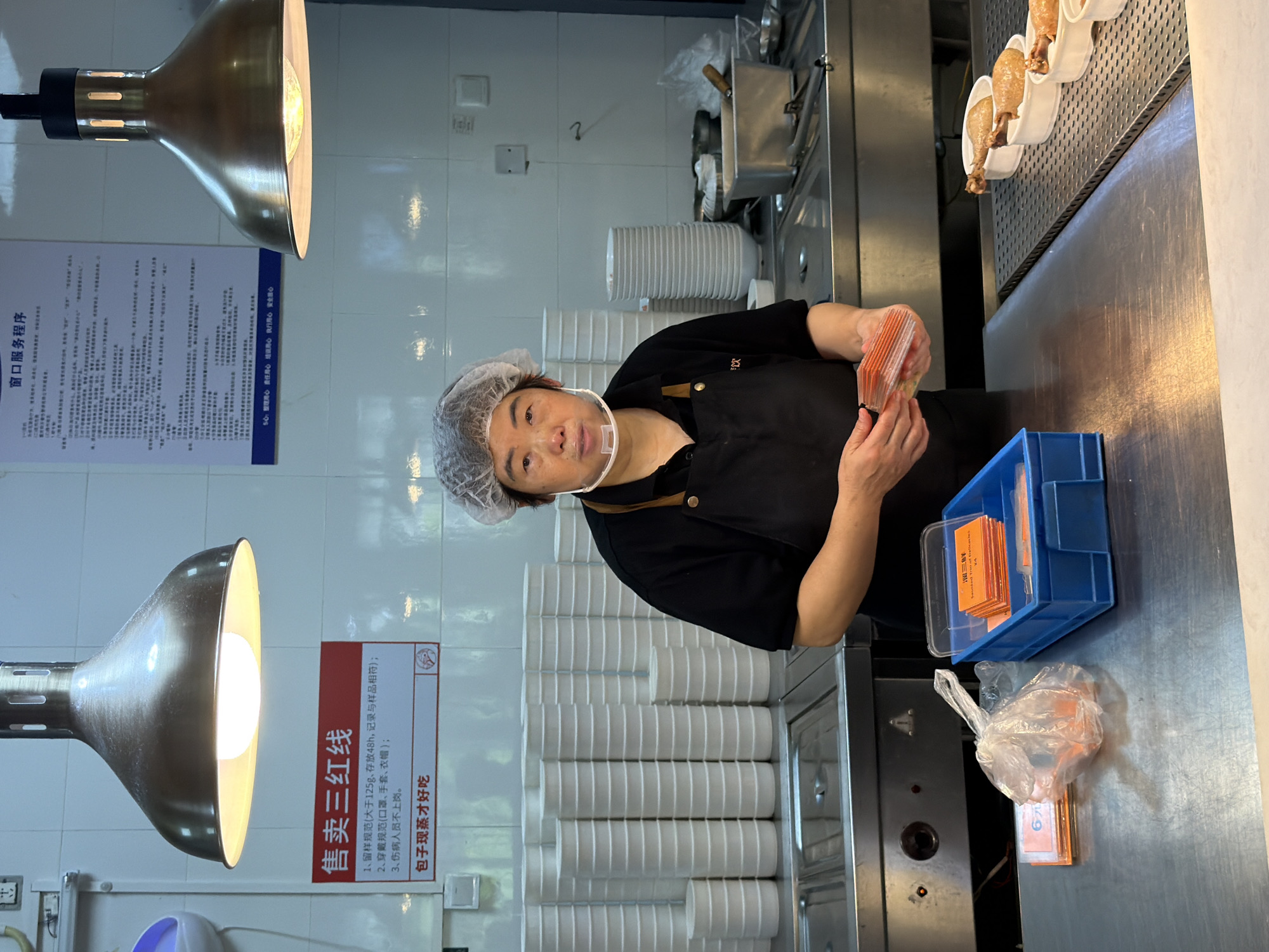 An indoor scene in what appears to be a commercial kitchen or food service counter in Jiangjin, China, shows a woman engaged in food preparation or service. The woman, who appears middle-aged, wears a black short-sleeved top, a black apron, a hairnet, and a transparent face shield, indicating adherence to hygiene standards. She is looking towards the viewer while holding a stack of small, orange rectangular items, possibly labels or price tags, in her hands.
The counter in front of her is stainless steel, and a blue plastic crate filled with similar orange cards is visible. Next to the crate is a clear plastic bag containing more of these cards, one clearly marked "6元" (6 yuan). In the background, stacked white bowls or containers are neatly arranged on a shelf. To the right, on an elevated warming rack, several chicken drumsticks or similar poultry items are displayed in individual white holders, ready for serving. Two large, industrial-style metallic lamps hang overhead, brightly illuminating the workspace.
Two signs written in Chinese are affixed to the white tiled wall. The sign on the left, titled "窗口服务程序" (Window Service Procedure), lists operational guidelines, concluding with a "5心" (5 Hearts) motto emphasizing values like organization, responsibility, care, assurance, and safety. The red-bordered sign on the right, labeled "售卖三红线" (Three Red Lines for Selling), outlines food safety and hygiene regulations, including specifications for food sample retention, staff attire (masks, gloves, hats), and prohibiting sick employees from working. It also states, "包子现蒸才好吃" (Steamed buns are only delicious when freshly steamed), suggesting buns might be sold here. The overall impression is a clean, well-organized, and brightly lit food preparation environment.