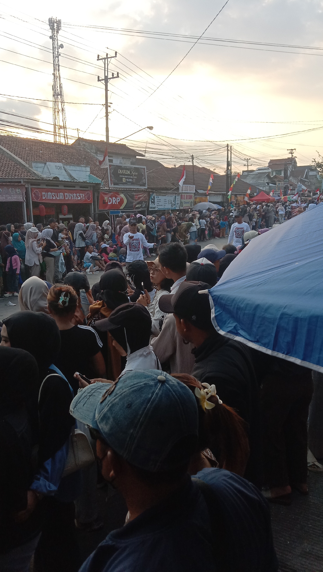 The image captures an outdoor gathering of people in what appears to be a street in the city of Pesantren, Indonesia, at dusk. The sky is overcast with hues of orange and yellow. In the background, there are buildings with tiled roofs, utility poles with numerous wires, and a tall telecommunications tower. Several signs are visible, including one that reads "DIMSUM DINASTI" and another for "Djarum Super" which is a brand of Indonesian kretek cigarettes. Indonesian flags are displayed on some of the buildings.
The foreground and midground are filled with a dense crowd of individuals, suggesting an event or festival. Many people are wearing casual clothing, and some are wearing headscarves, indicating a culturally diverse or predominantly Muslim population. A few individuals are standing and gesturing, possibly addressing the crowd or participating in an activity. Others are sitting on the ground, and many are standing and observing.
Notable objects include a large blue umbrella covering a portion of the right side of the frame, potentially providing shade or shelter. The lighting suggests it is late afternoon or early evening, with the sun casting a warm glow. The overall atmosphere seems to be one of a community event, with a significant turnout of people.