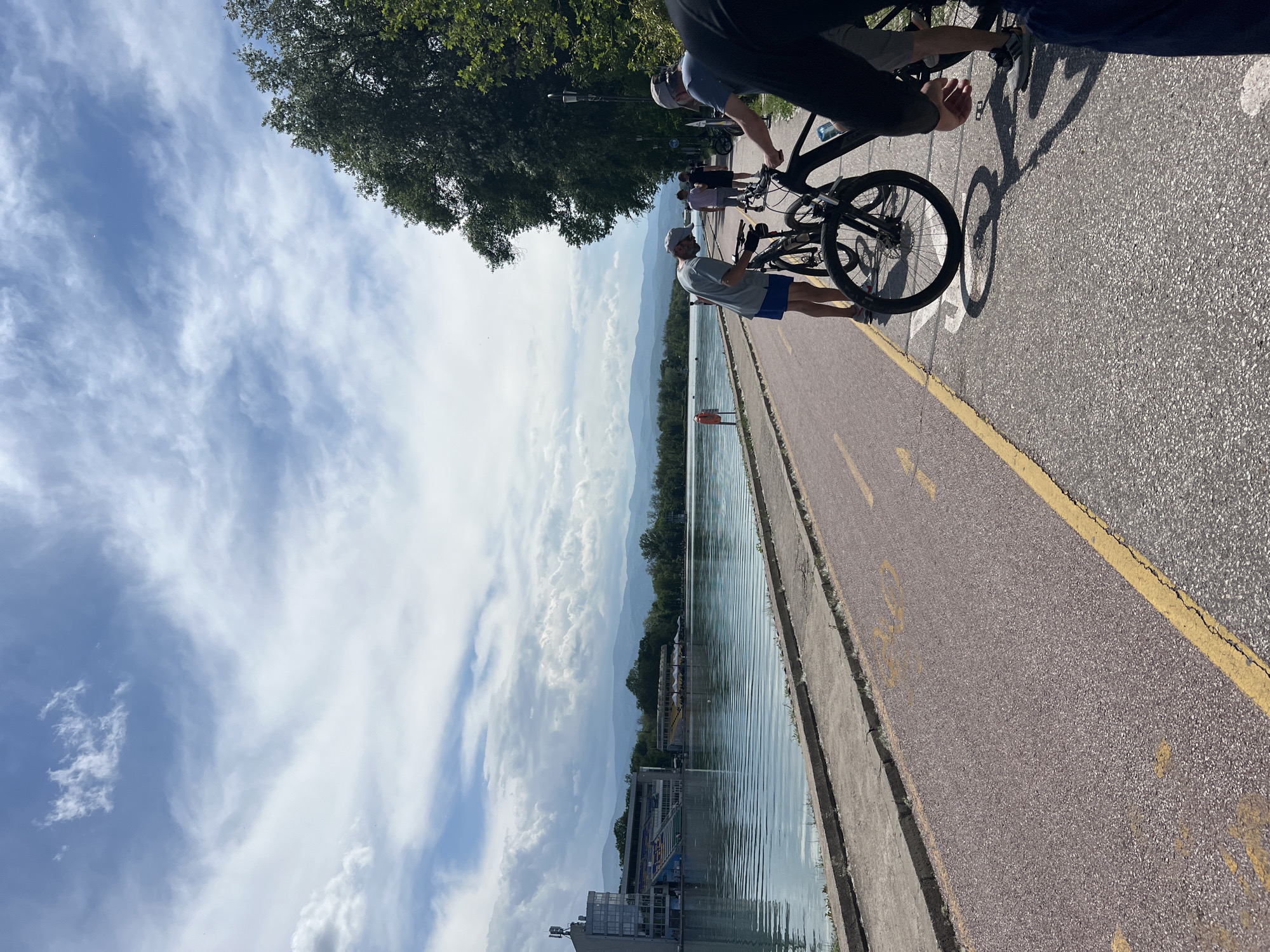 The image depicts a scene along a paved walkway next to a lake or wide river. People are present, some with bicycles. A man wearing a hat, gray shirt, and blue shorts is standing near the edge of the walkway, looking out over the water. Another person is seen partially, standing with a bicycle. A yellow line marks the edge of the walkway. Trees are visible on the left. Across the water, there is a building with a ramp, and distant mountains are visible. The sky is partly cloudy. It is most likely near the Plovdiv Rowing Channel in Bulgaria.