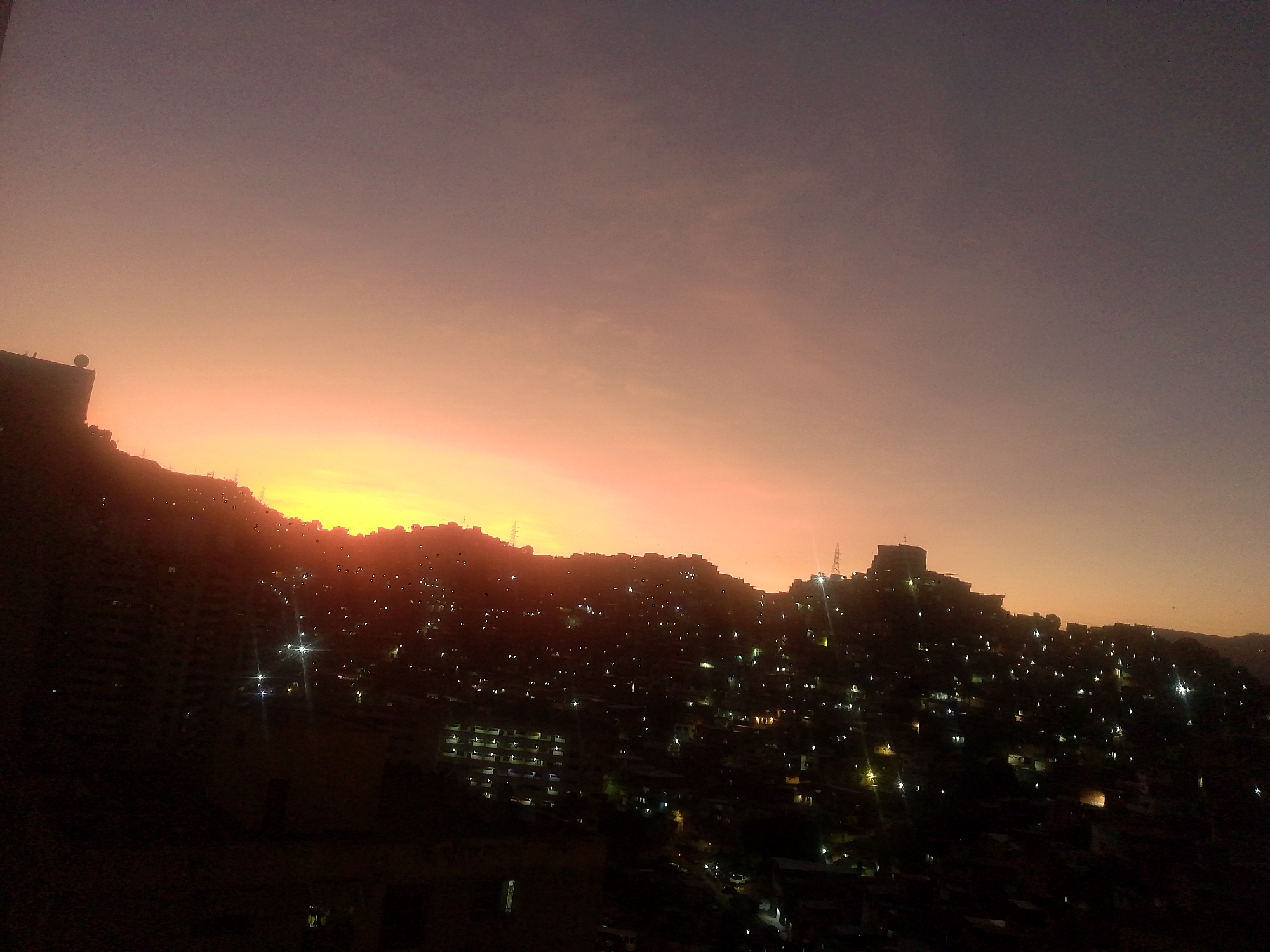 The image depicts a sunset over Caracas, Venezuela, showing a hillside densely populated with buildings illuminated by scattered lights. The sky transitions from a bright orange/yellow horizon to a dark purplish hue above. The lower portion of the image is dark, obscuring details in the foreground, while the hilltop houses are sharply defined against the glowing sky. Electrical towers are faintly visible on the hilltop. There is no visible evidence of people or other objects.
