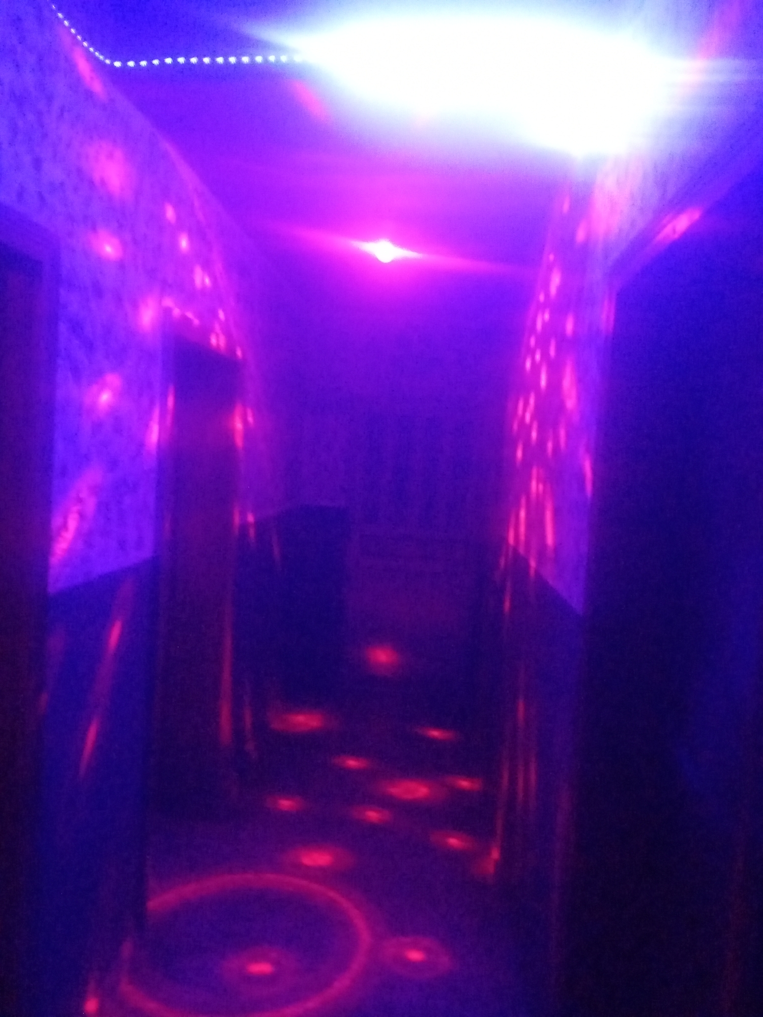 The image depicts an indoor environment, likely a hallway or corridor, characterized by intense purple and blue lighting. The walls are illuminated with scattered pinkish-red spots, suggesting a light effect or projections. A bright white light emanates from the ceiling at the far end of the corridor, casting a strong glow. Red circular patterns and smaller red spots are visible on the floor, possibly from lighting fixtures or projections. The overall scene conveys a vibrant, possibly playful or entertainment-oriented atmosphere. No people or specific actions are discernible. The provided location information, "Sabon Gari Nassarawa, Nigeria," is not visually supported by any elements within the image itself.