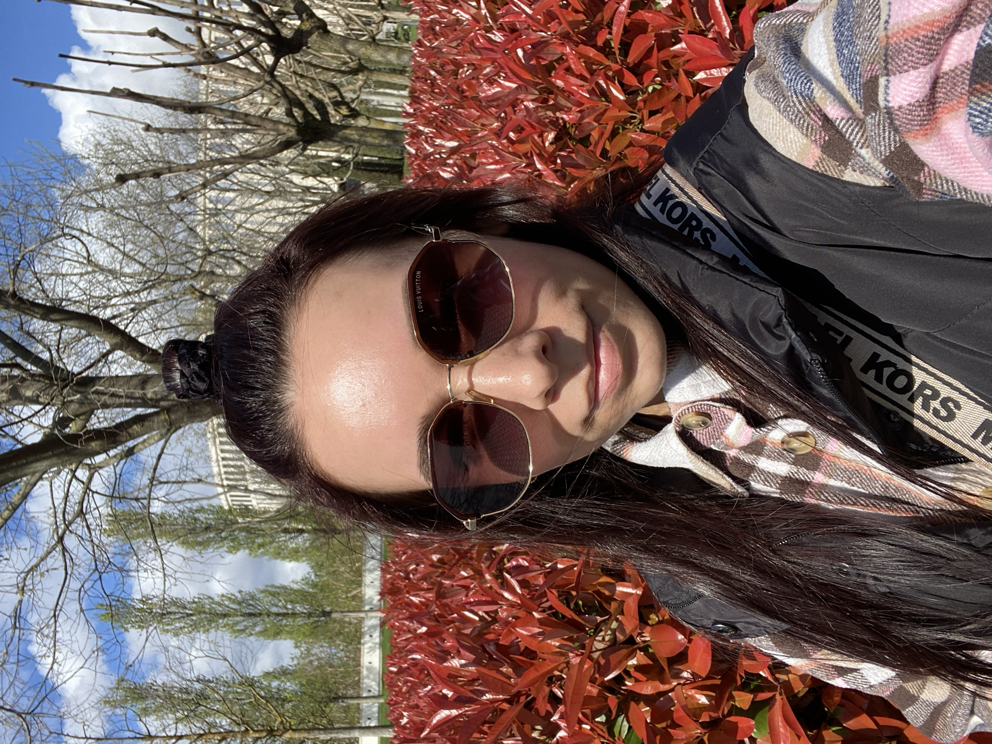 A woman poses for a selfie, wearing sunglasses marked with the name "LOUIS VUITTON," a black jacket with "MICHAEL KORS" printed on the straps, and a plaid shirt. Her dark hair is pulled back in a bun. The background features a hedge of red-leaved plants, trees with bare branches against a blue sky with white clouds, and a building in the distance. The image does not provide enough information to pinpoint the specific location in Krasnodar, Russia.