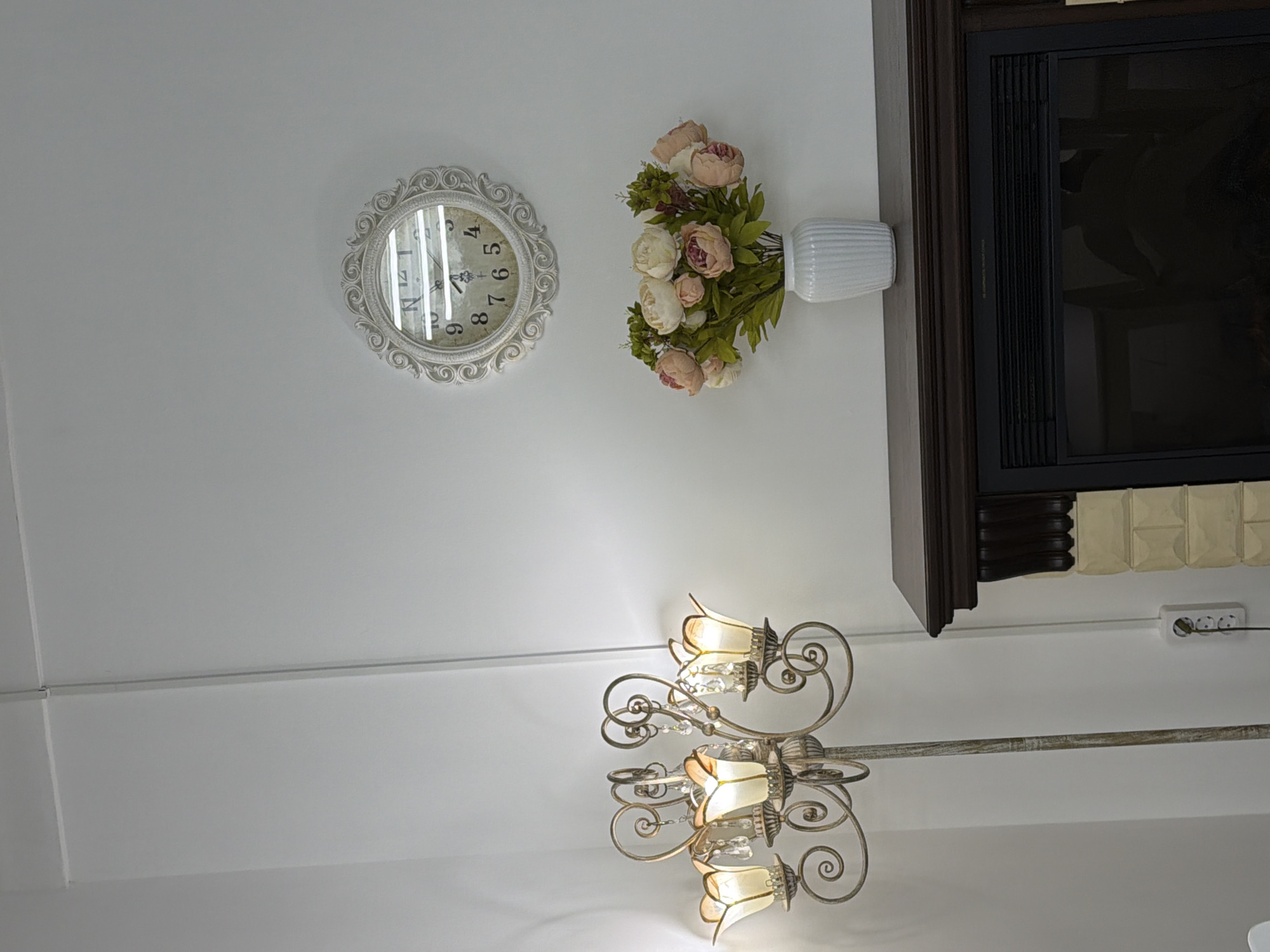 An interior shot depicts a decorative room with a white wall and ceiling. On the wall, an ornate white-framed clock with Roman numerals is visible, though reflections obscure most of the face, showing numbers like "4", "6", "8", and "9". To the right of the clock, a white ribbed vase holds a bouquet of artificial flowers, primarily pink and cream-colored peonies or roses with green foliage. Further to the right and above the flowers, a dark wooden structure, possibly a mantelpiece or part of a built-in unit, frames a dark, rectangular screen, likely an electric fireplace or television.
Below the wooden structure, a white power strip with three outlets is plugged into the wall, with a white cable extending horizontally across the white wall. Hanging from the ceiling in the lower part of the frame is a large, elaborate chandelier with a distressed white or antique gold finish, featuring multiple light fixtures shaped like inverted flowers and adorned with dangling crystal accents. The lights on the chandelier are illuminated, casting a warm glow. No people are present, and no specific activity is taking place. The setting appears to be a decorated living space. No explicit location cues other than the interior decor are present.