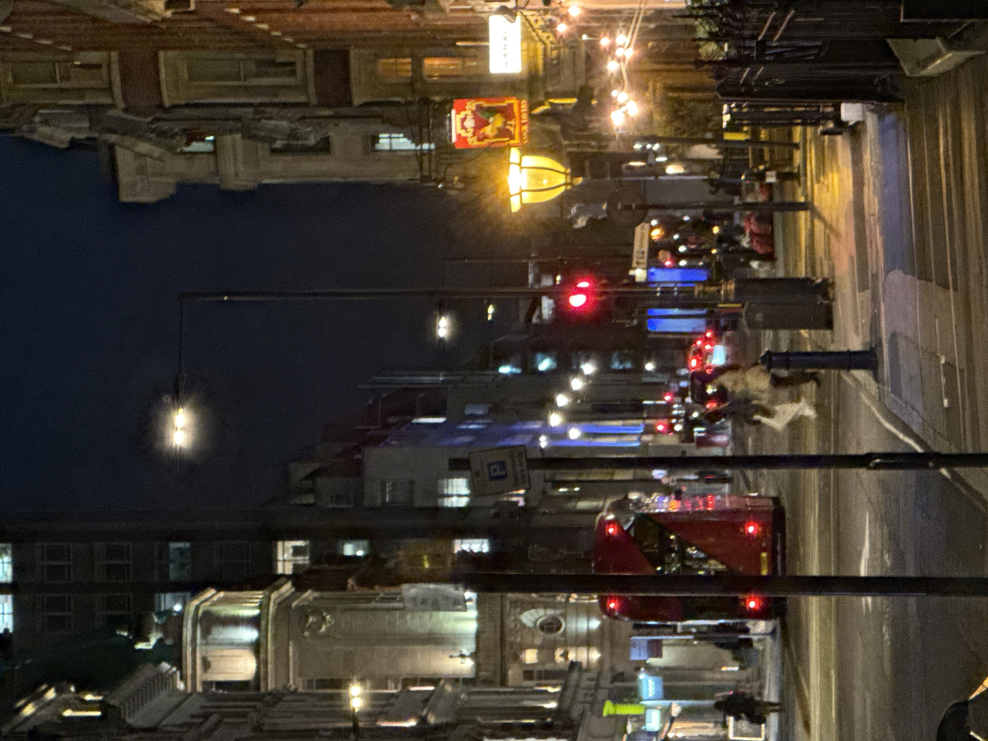 The image captures a vibrant city street scene at night, taken from an elevated vantage point, likely in London, United Kingdom. The street is bustling with activity, illuminated by a myriad of artificial lights against a dark night sky.
Tall, multi-story buildings line both sides of the street, their windows and ground-floor establishments casting various glows onto the pavement. The road is busy with traffic; a prominent red double-decker bus, a characteristic feature of London, is seen in the mid-ground, moving away from the viewer, its red taillights glowing. Other vehicles are also visible, indicated by the red streaks of their taillights and the occasional white glint of headlights. A red traffic light is clearly visible on the right side of the street, suggesting vehicles are stopped or slowing down.
Pedestrians are present on the sidewalks and possibly crossing the street, appearing as blurred figures due to movement or the low-light conditions. The street is adorned with typical urban furniture, including tall lamp posts, which emit bright flares of light.
Several illuminated signs add to the nocturnal ambiance. A bright yellow, possibly bell-shaped, illuminated lamp or sign hangs over the street on the left. Nearby, a rectangular red sign features a colorful illustration, resembling a pub sign or advertisement. Above this, a white rectangular sign has some indistinct text, possibly "MAHAC". Further down the street, a blue square sign with a white "P" symbol (indicating parking) is visible above the road. The overall scene depicts a lively and busy urban environment after dark.