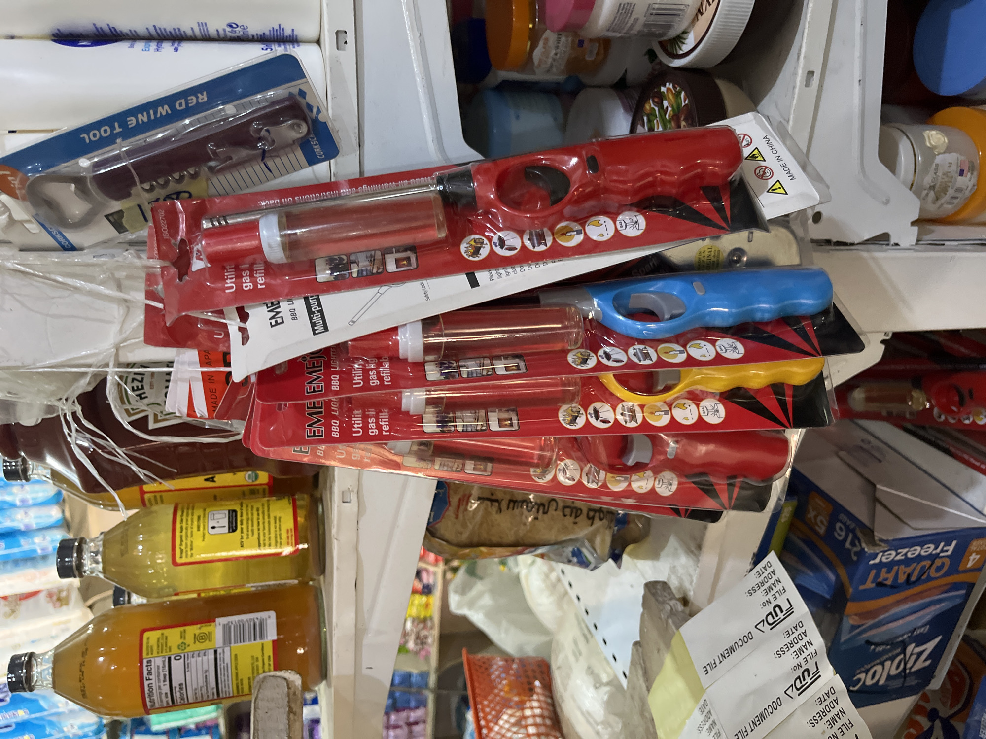 The image depicts a shelf in a store in Bukuru, Nigeria, displaying various household items. In the foreground, several brightly colored utility lighters are packaged and arranged vertically. These lighters are predominantly red, with some blue and yellow variations also visible. Each lighter's packaging includes graphic illustrations depicting their intended uses, such as lighting grills or stoves. Behind the lighters, a red bottle, possibly ketchup or a similar condiment, stands prominently. Further back, on the same shelf, are bottles of what appear to be vinegar or cooking oil, with yellow labels. A red plastic basket is visible to the left, and in the background, shelves are stocked with an assortment of goods, including what looks like packaged food items and cleaning supplies. The overall environment suggests a retail setting with a variety of products for sale.