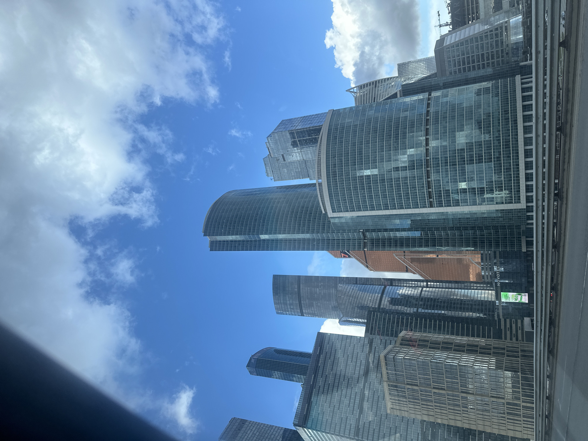 The image depicts a low-angle view of the Moscow International Business Center (Moscow-City) skyline, seen from an elevated position, potentially from a bridge or high-rise building. Numerous skyscrapers with glass facades dominate the scene, reflecting the blue sky and scattered clouds. The sky is visible between the buildings. The overall architectural style is modern, with some buildings featuring curved designs. A part of a concrete barrier is visible on the right side, suggesting the viewpoint is from a road or a bridge. There is a hint of construction work visible at the top right.