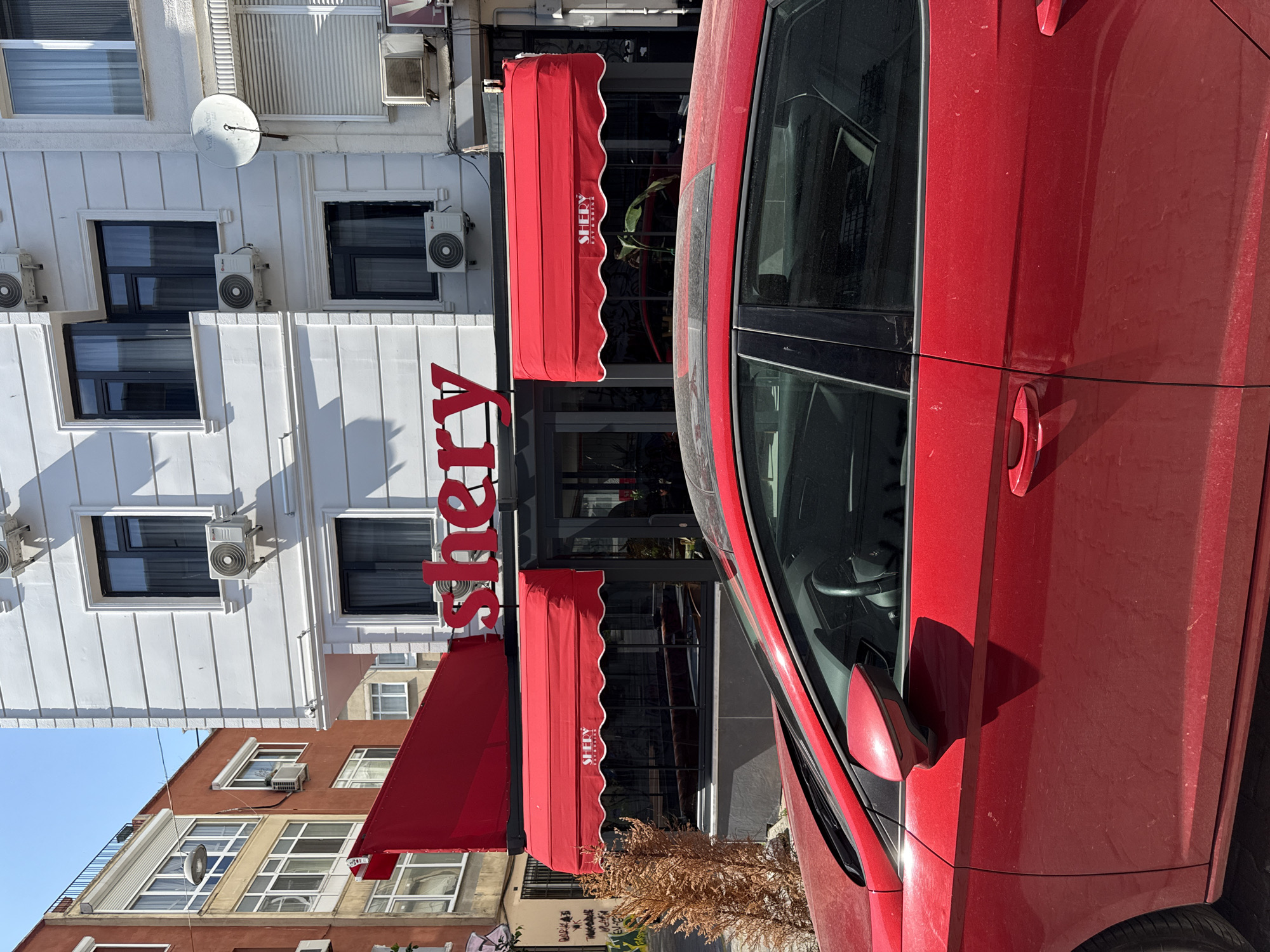 The image depicts a street scene in Kadıköy, Turkey, featuring a red car parked in front of a building. The building has a white facade with several windows and air conditioning units. A red awning extends over the ground floor of the building, displaying the name "SHERY" in large, red, capitalized letters. Below the awning, the windows of what appears to be a cafe or restaurant are visible, with seating and plants inside. To the left of the building, another building with an orange facade is partially visible. A dry, brown shrub is located to the left of the entrance. The sky is clear and blue. The red car in the foreground is dusty, showing signs of wear and tear. The overall impression is a snapshot of an urban environment on a sunny day, highlighting the facade of a business and its surroundings.