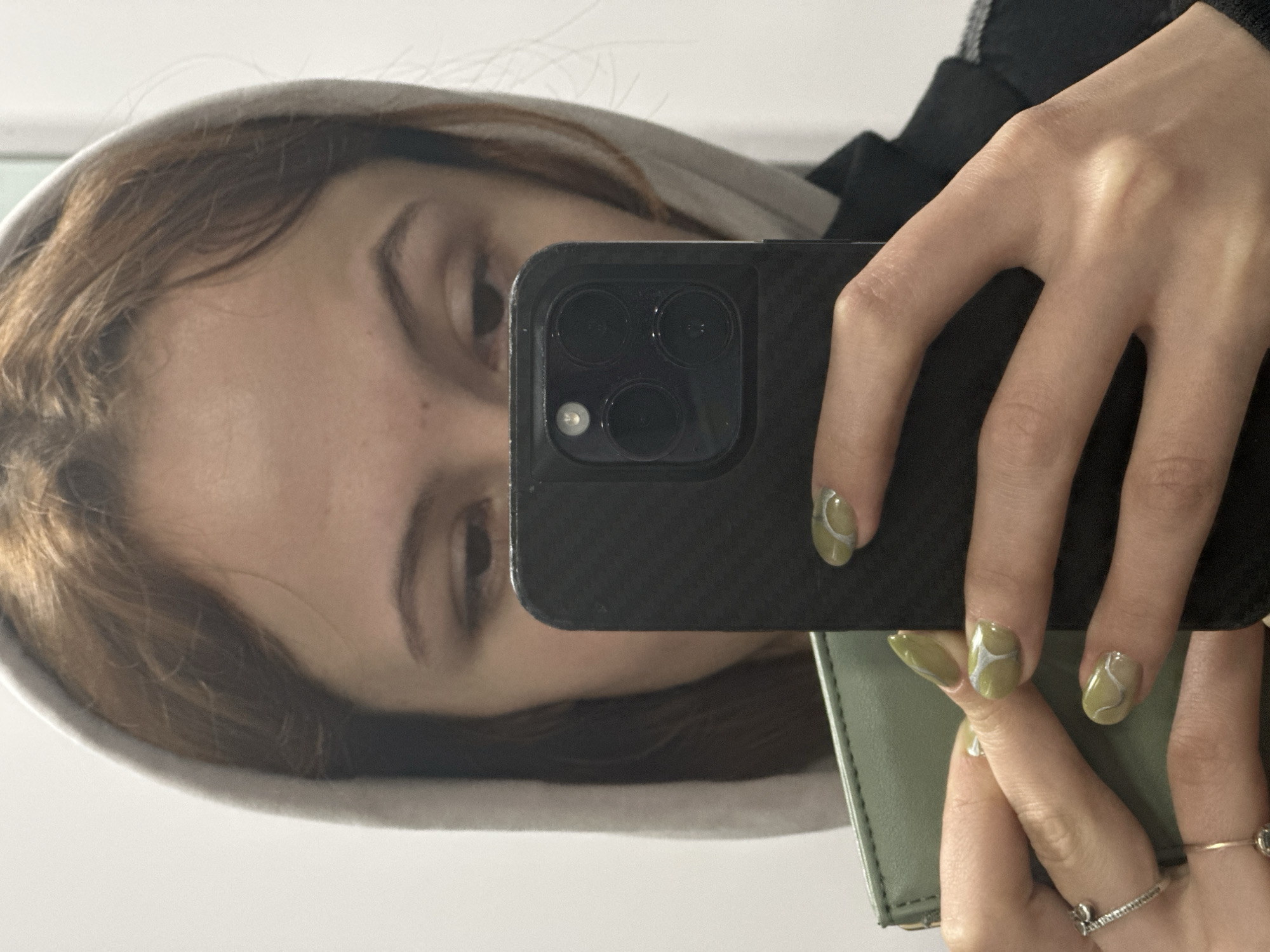 A person is taking a selfie with a black smartphone in front of a mirror. The person is wearing a light gray hoodie and a black jacket. The person's nails are painted with a light green color and silver swirls. The person is also holding a light green wallet. The location cannot be determined based on this image.