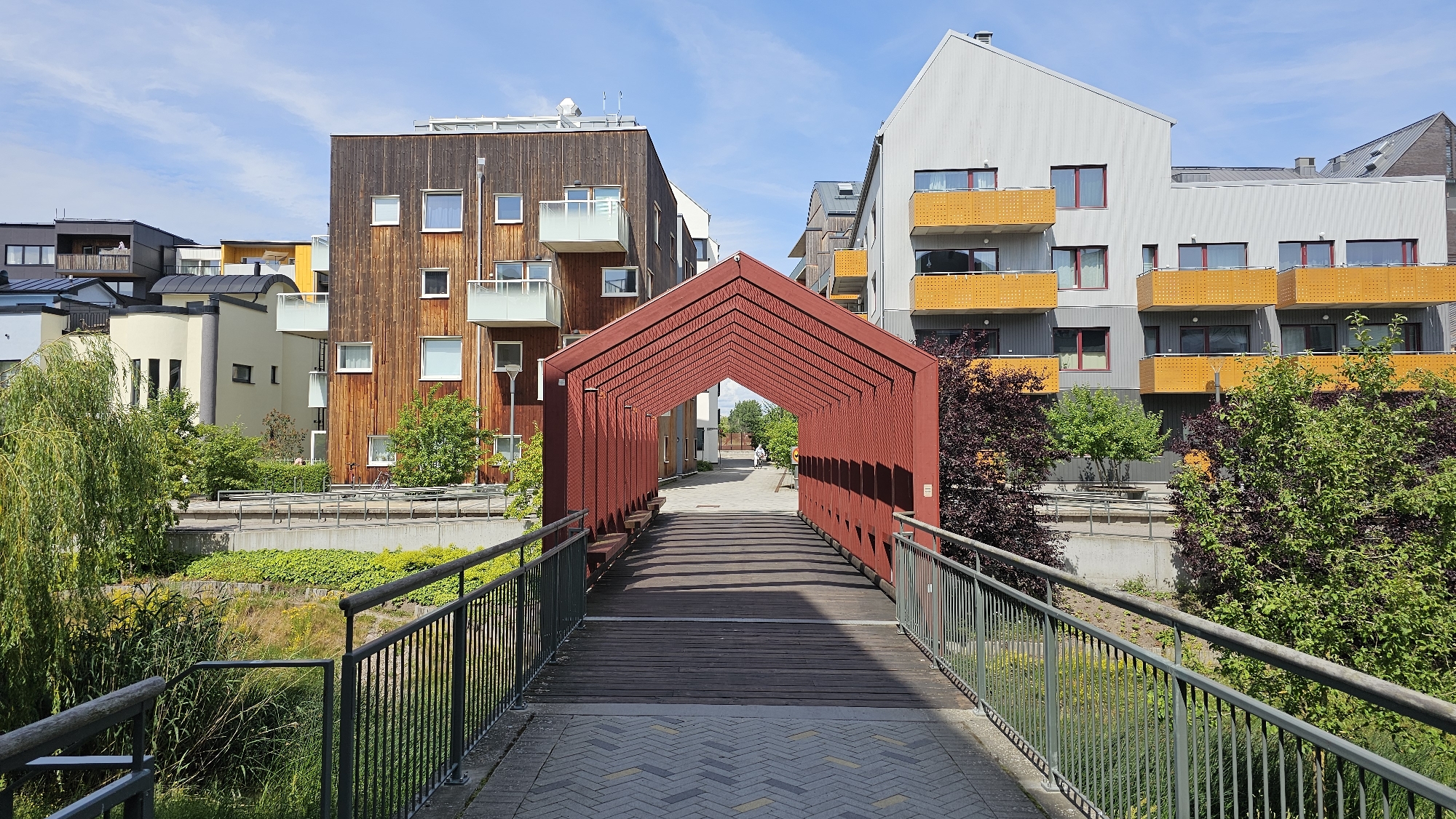 The image shows a pedestrian bridge leading to a modern residential area in Linköping, Sweden. The bridge has a red, arched canopy supported by a metal frame, and its wooden walkway is lined with metal railings. Beyond the bridge, contemporary apartment buildings with varied facades and balconies are visible. Some buildings feature dark wood paneling, while others are clad in grey and white materials with vibrant yellow balconies. Green trees and lush vegetation surround the buildings, creating a pleasant urban environment. The sky is blue with scattered clouds, indicating fair weather. In the distance, a person can be seen walking along a paved path. The overall scene depicts a well-designed, modern living space integrated with nature in a Scandinavian city.