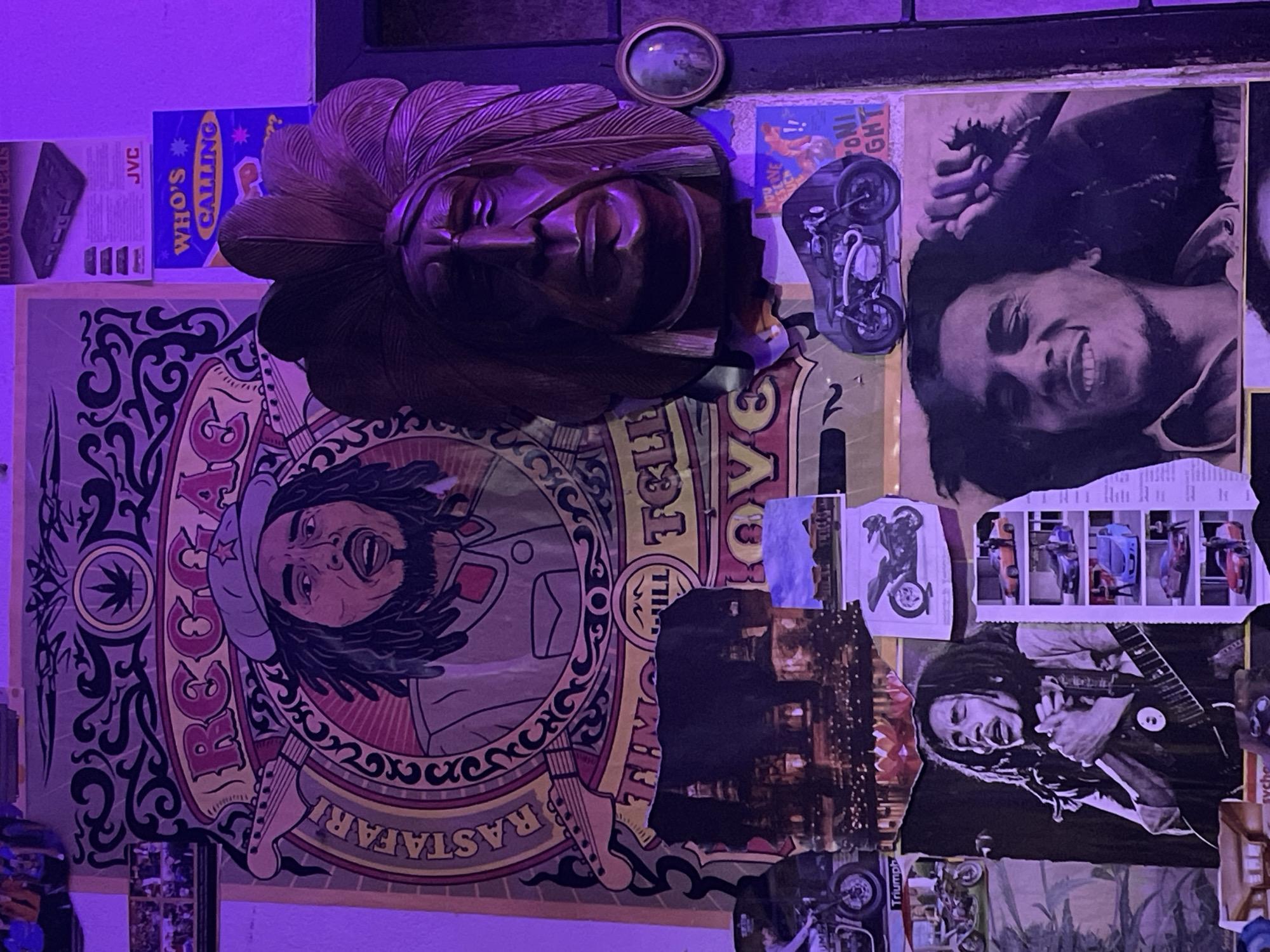 The image depicts a wall covered in a collage of posters, artwork, and a wooden carving. The dominant theme appears to be reggae music and its associated culture, with multiple images of Bob Marley visible. There are also posters with text related to "Reggae" and "Rastafari." A prominent wooden carving of a Native American chief wearing a feathered headdress is mounted on the wall. The lighting is predominantly purple, suggesting the presence of mood lighting or a specific light source. The overall impression is that of a decorated interior space, possibly a room or a bar, adorned with thematic decorations. The provided context mentions the location as Chalong, Thailand, which implies this is a scene from a venue in that city. No people are actively present or interacting in the scene, only static representations on posters and artwork. The environment appears to be indoors, with the purple lighting creating a distinct atmosphere.