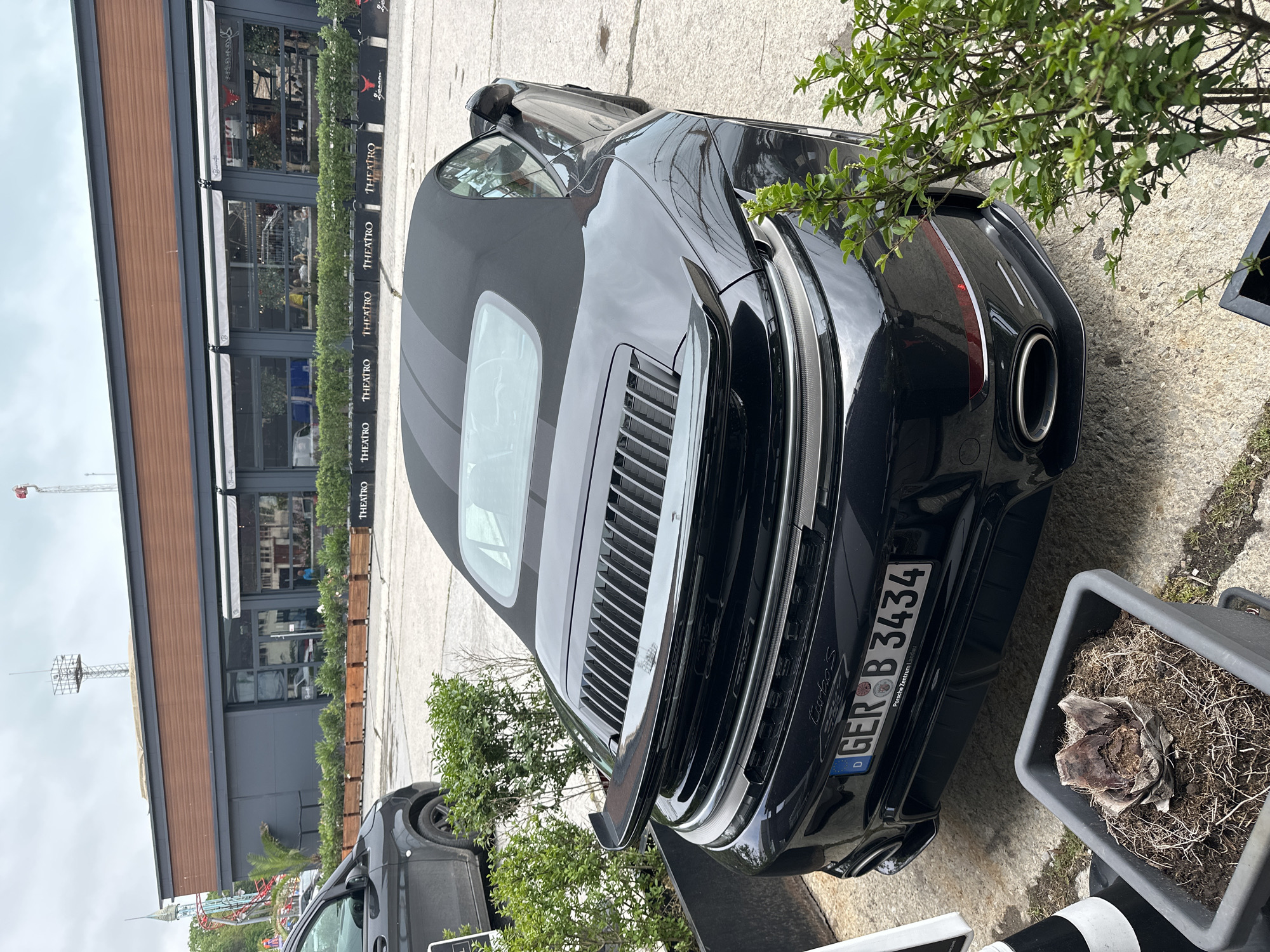 An overhead view, rotated 90 degrees clockwise, captures the rear three-quarter of a black Porsche 911 Turbo S Cabriolet parked on a paved surface. The car, with its soft top up, features a distinctive rear design with horizontal engine cover slats, integrated spoiler, and dual exhaust pipes. The German license plate, "GER B 3434," is clearly visible, with "Porsche Zentrum Lübeck" inscribed on its frame.
To the left of the car is a modern building with large glass windows, suggesting a restaurant or cafe. Several white awnings are partially extended above the windows. "THEATRO" is repeatedly displayed on dark dividers or planters lining the facade, and the word "Brownie" can also be seen on an awning. Through the glass, some interior details and possibly people are discernible.
The ground is concrete or asphalt. Various green shrubs and small trees are planted along the side of the building and near the parked car. A large, grey planter with a dead or dormant plant stands next to the car's rear. In the far background, under an overcast sky, parts of a tall antenna or construction crane are visible, along with what appears to be part of a roller coaster or amusement park ride. The setting suggests a commercial or leisure area.