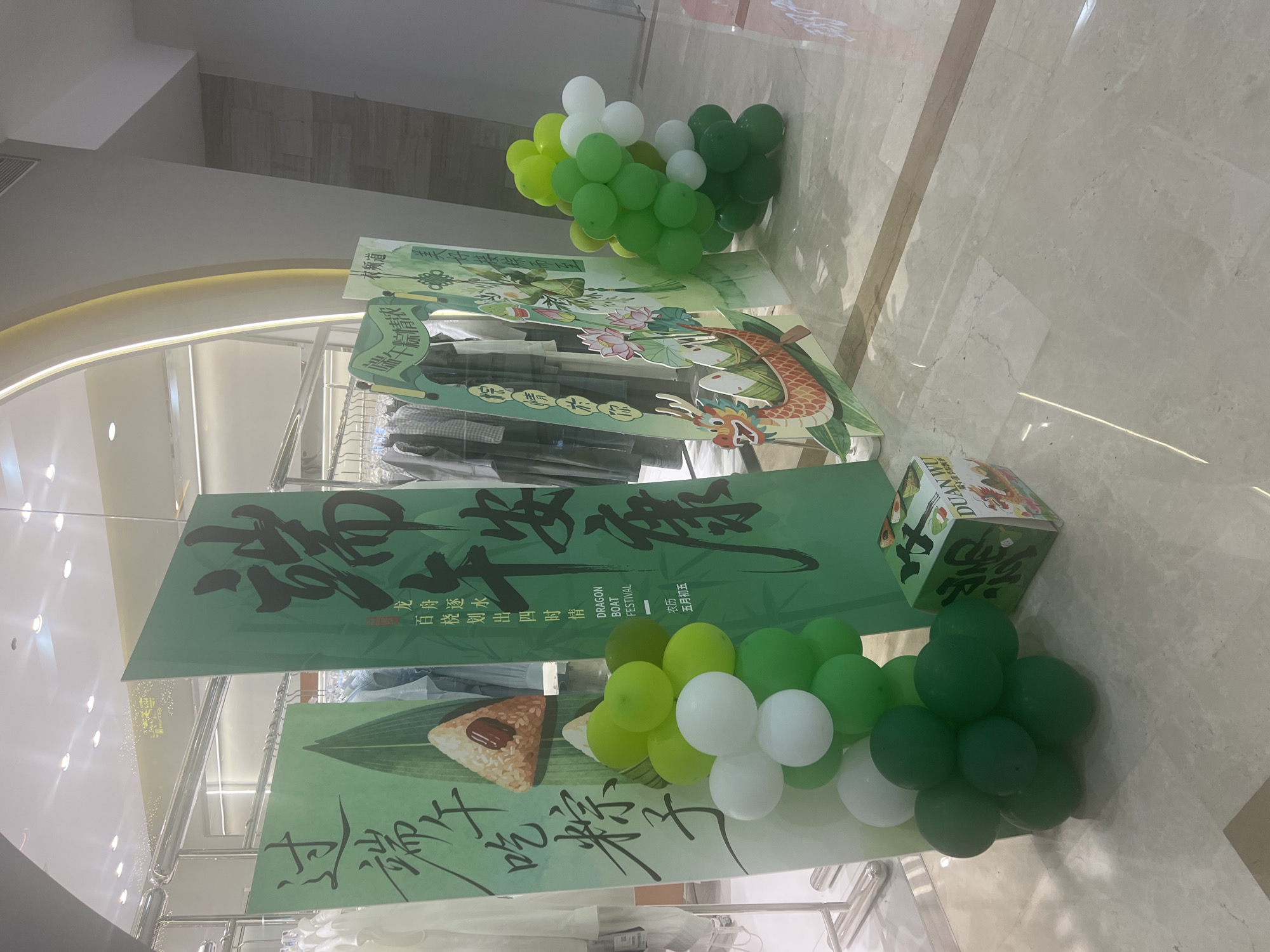 The image depicts a retail store display for the Dragon Boat Festival, situated in Liandu District, China. The foreground features clusters of green and white balloons arranged on the floor. To the left, a large green display board with Chinese calligraphy and an illustration of a zongzi (rice dumpling) is visible. Next to it, another green display board features more Chinese calligraphy and a stylized dragon boat with lotus flowers. A smaller green display board further right shows similar festive imagery. In the center, a square cardboard box with "DUAN WU" and Chinese characters is placed on the floor, adorned with illustrations related to the festival. The background shows racks of clothing within the store, illuminated by overhead lights. The floor is a polished marble finish.