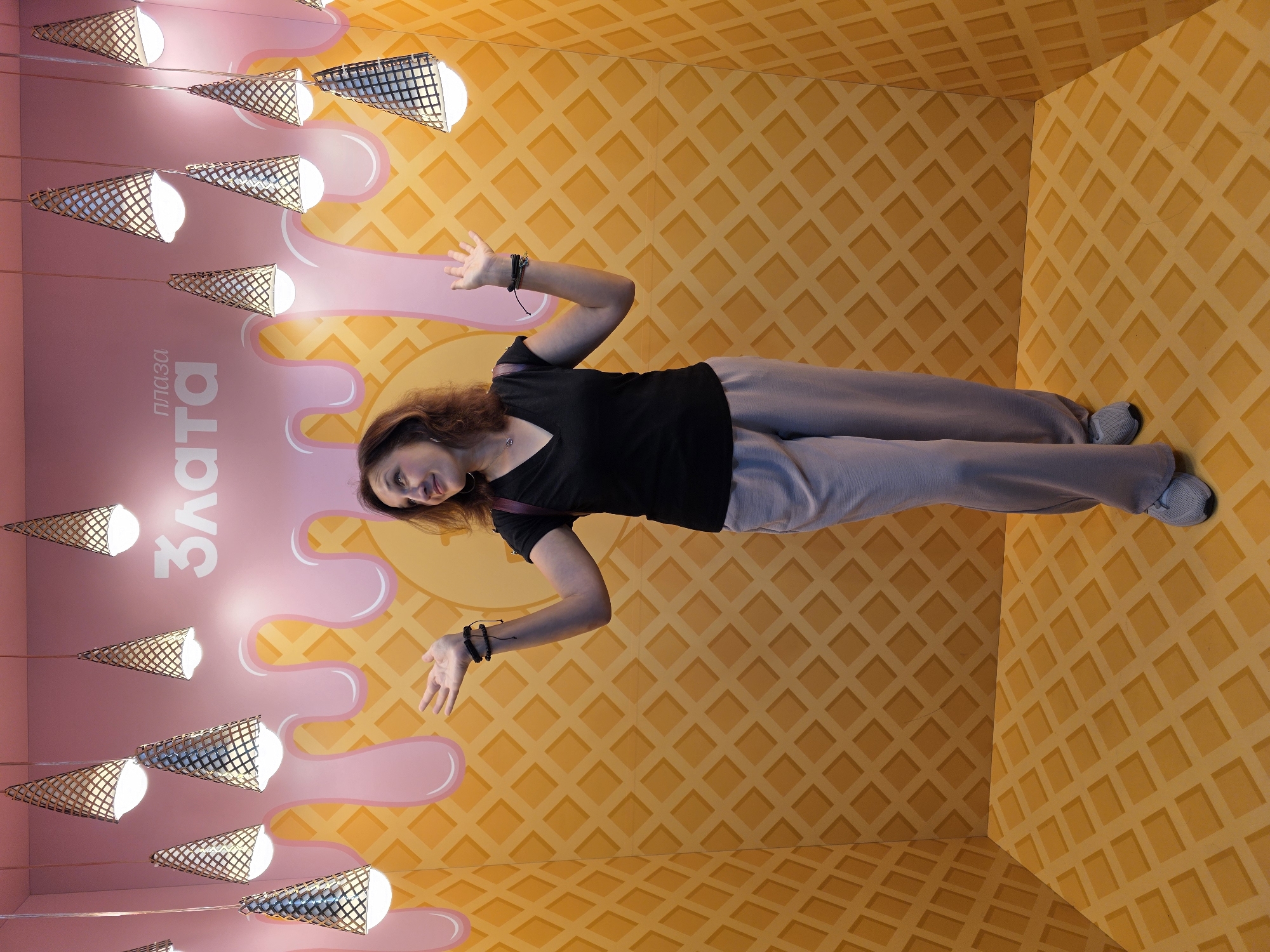 The image depicts a woman posing in an indoor photo zone designed to resemble a waffle cone with dripping pink frosting. The walls and floor are covered in a yellow waffle pattern. Overhead, multiple cone-shaped lamps hang, resembling ice cream cones. The woman, wearing a black V-neck shirt and grey pants, stands with her arms outstretched and hands open, looking up and to her right. On the wall behind her, in white lettering, is the text "Плаза Злата". This photo zone is located in the Plaza Zlata shopping center in Rivne, Ukraine.