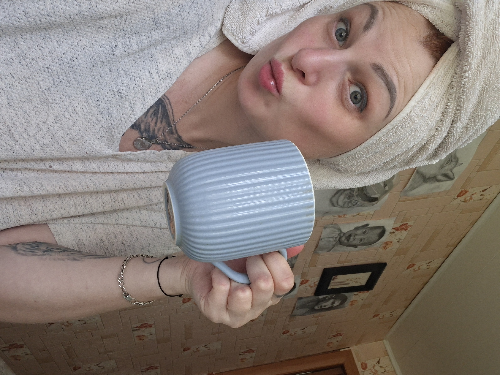 A young Caucasian woman with red hair is captured in a close-up, selfie-style shot, holding a light blue ribbed mug. She is wearing a light-colored, textured top and a white towel is wrapped around her head, covering her hair. Her lips are slightly parted, and she appears to be making a kissing or puckering expression. A silver necklace with a pendant is visible around her neck, and she has a tattoo on her chest. A black cord bracelet is on her wrist. The background consists of a pinkish-beige patterned wallpaper with framed drawings or photographs of people and animals. The scene is indoors, likely in a residential setting. The lighting suggests daytime. There is no specific information to indicate the location in the city of Myadzyel, Belarus.