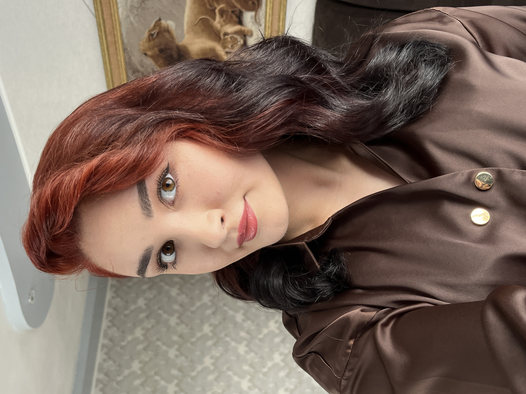 A young woman with red and black ombre hair is posing for a selfie indoors. She is wearing a brown satin shirt with gold buttons. Behind her, a painting of lions can be seen on the wall, and there is a patterned wallpaper. The ceiling has an unusual curved design with a light fixture. The image appears to have been taken in a residential setting in Starobaltachevo, Russia.