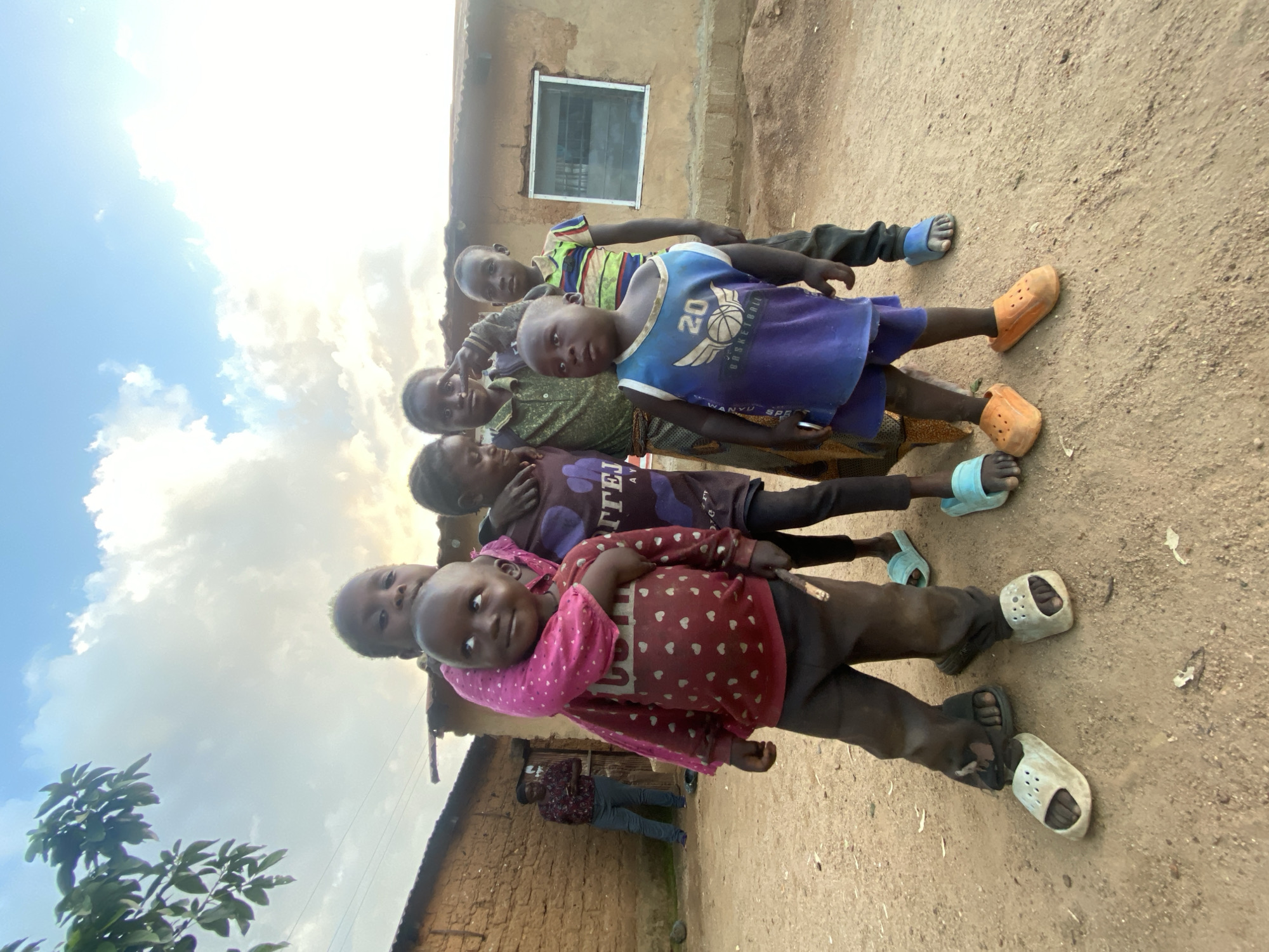 The image depicts a group of at least seven children standing closely together in an outdoor environment. The location is indicated as Riyom, Nigeria. The children are arranged in a line, partially obscured by each other. They are wearing casual clothing, including t-shirts, pants, and open sandals (Crocs). A building constructed of earthen materials is visible in the background, along with a man who is standing and is partially visible behind the children. The ground is dusty and there are clouds in the sky.